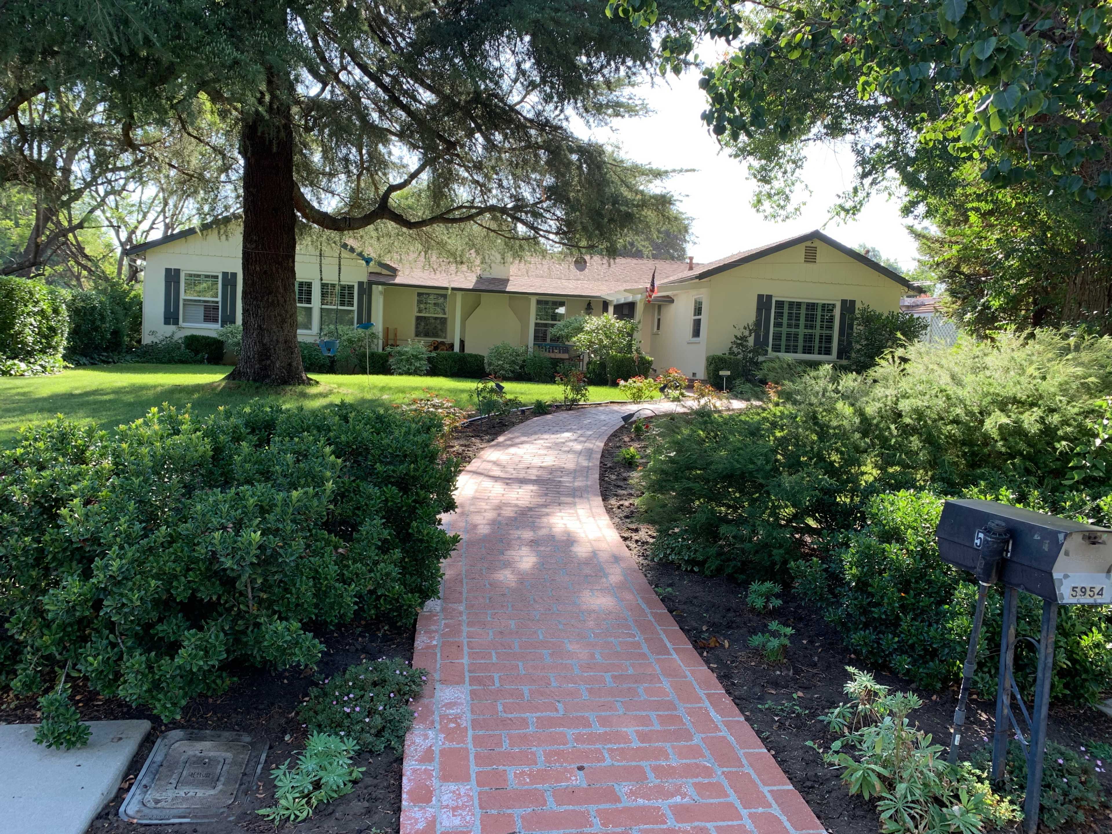 A brick pathway leads to a single-story house surrounded by well-maintained greenery.