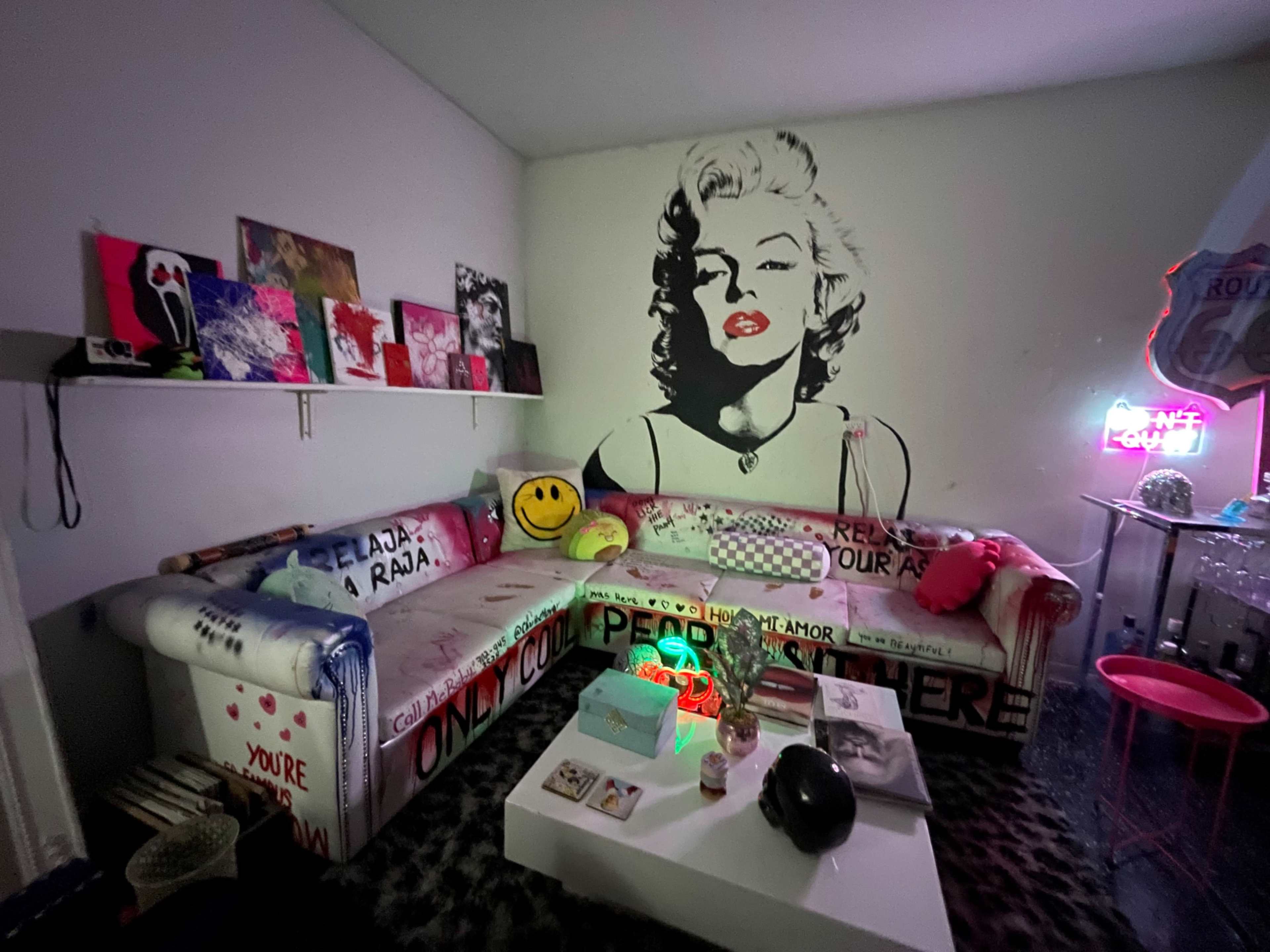 The image shows a cozy living room featuring a large sectional couch with vibrant, artistic decorations and a mural of Marilyn Monroe on the wall.