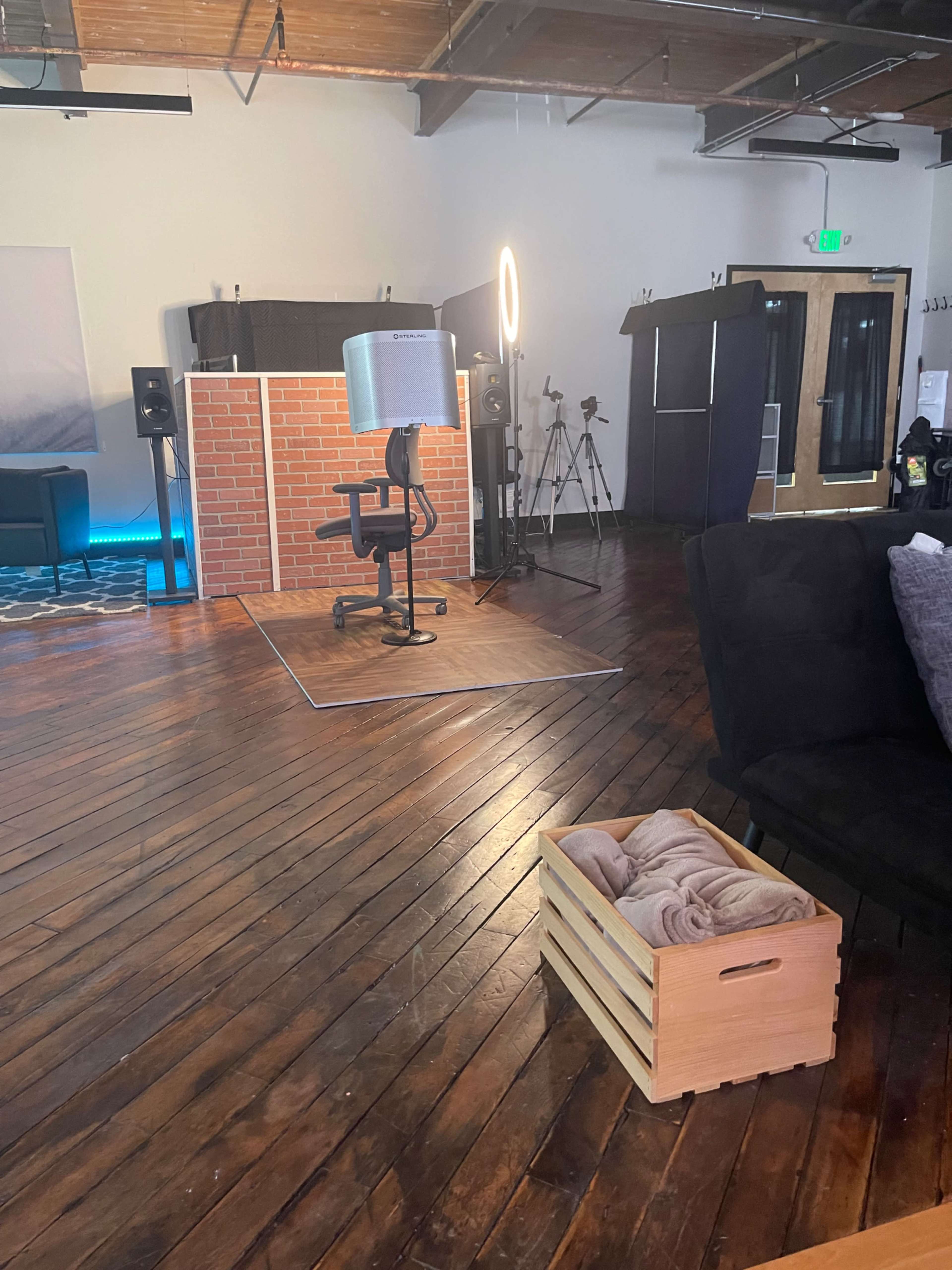 A spacious studio interior with a microphone setup on a swivel chair, a decorative brick backdrop, various photography equipment, and a wooden crate with blankets on the floor.