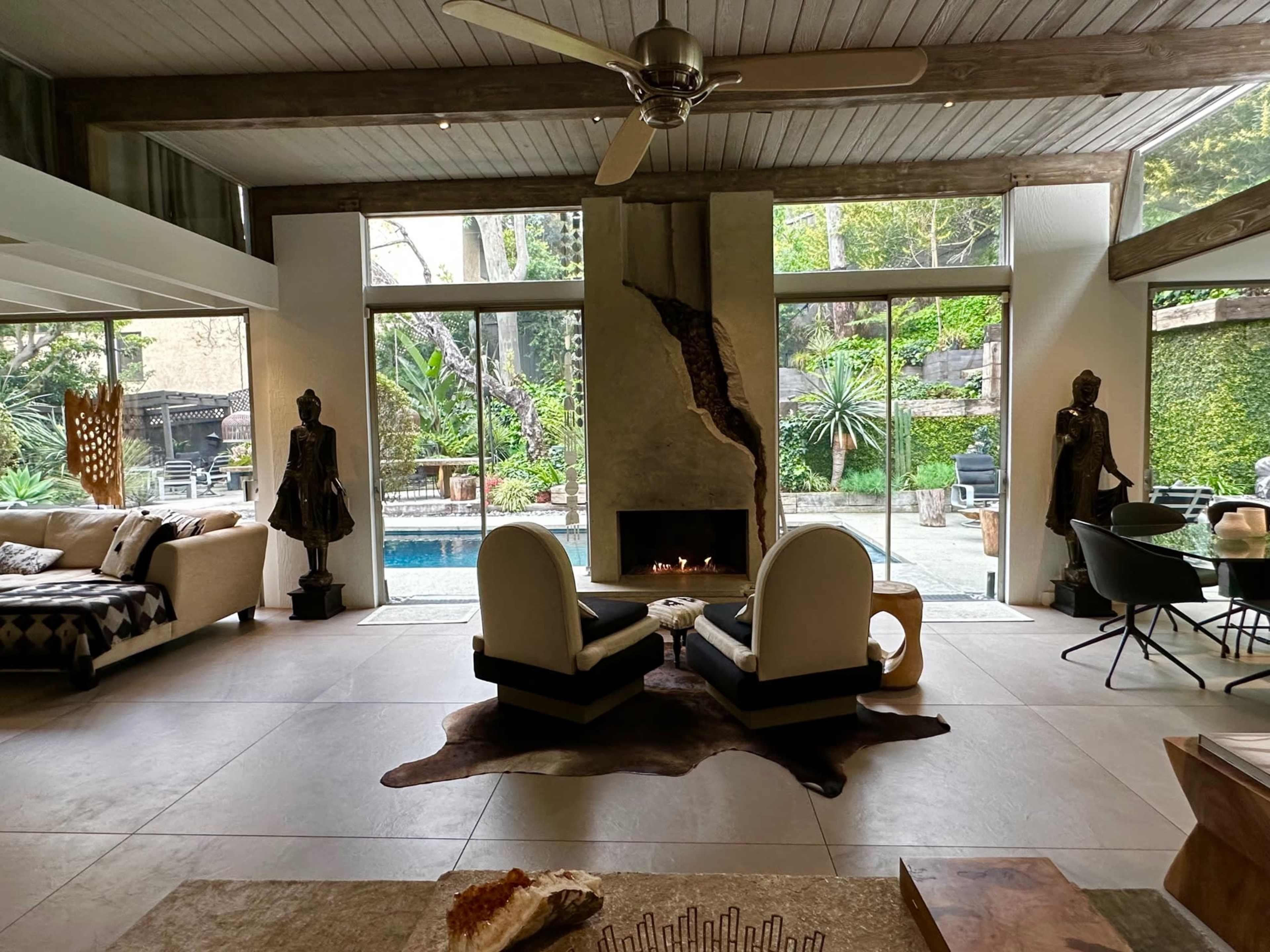 A modern living room with two white armchairs facing a fireplace, large windows revealing a pool and greenery outside, and various decorative elements throughout the space.