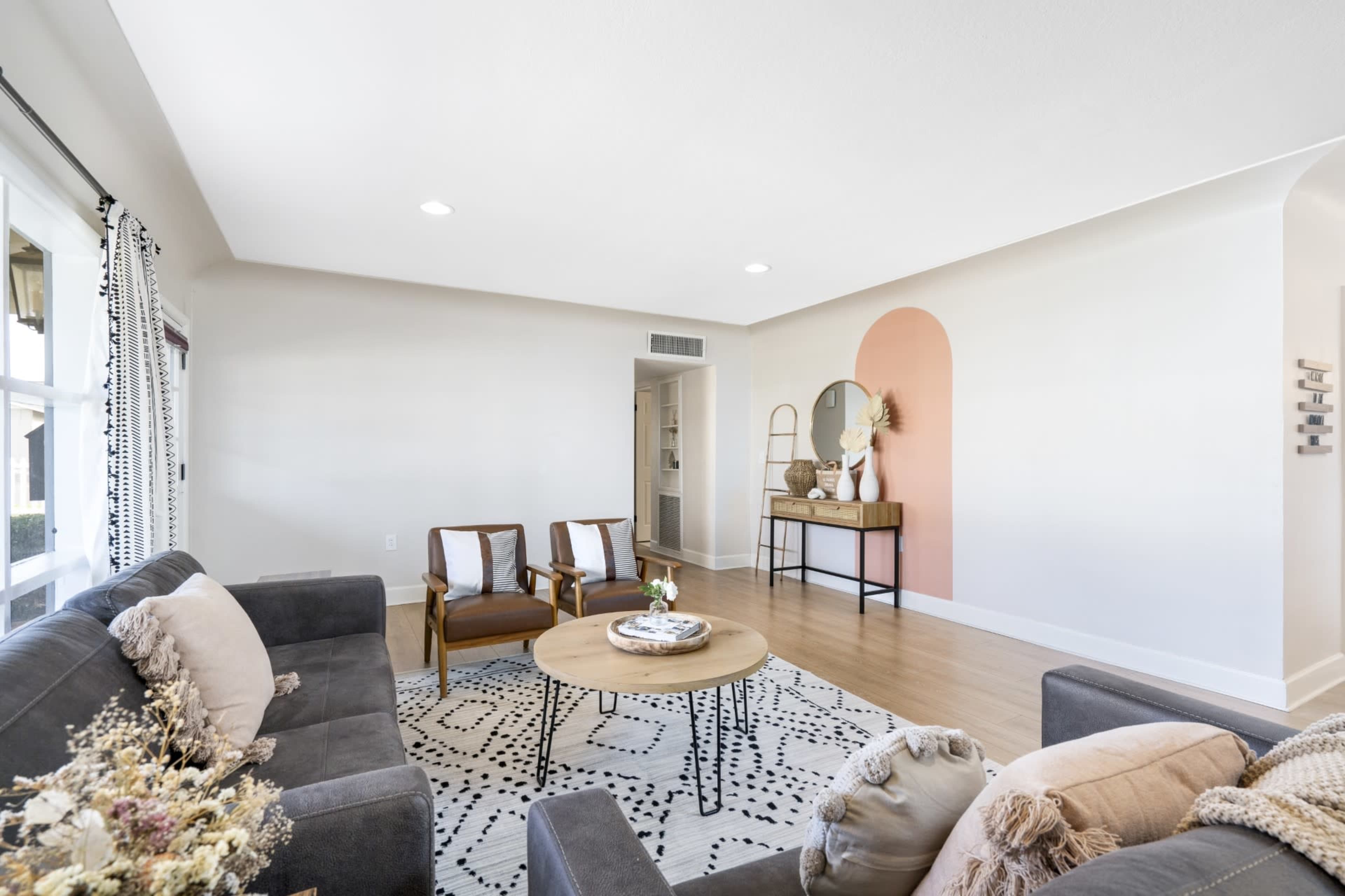 The image shows a modern living room featuring a gray couch, wooden chairs, a round coffee table, and a decorative console against a pale wall with a coral accent.