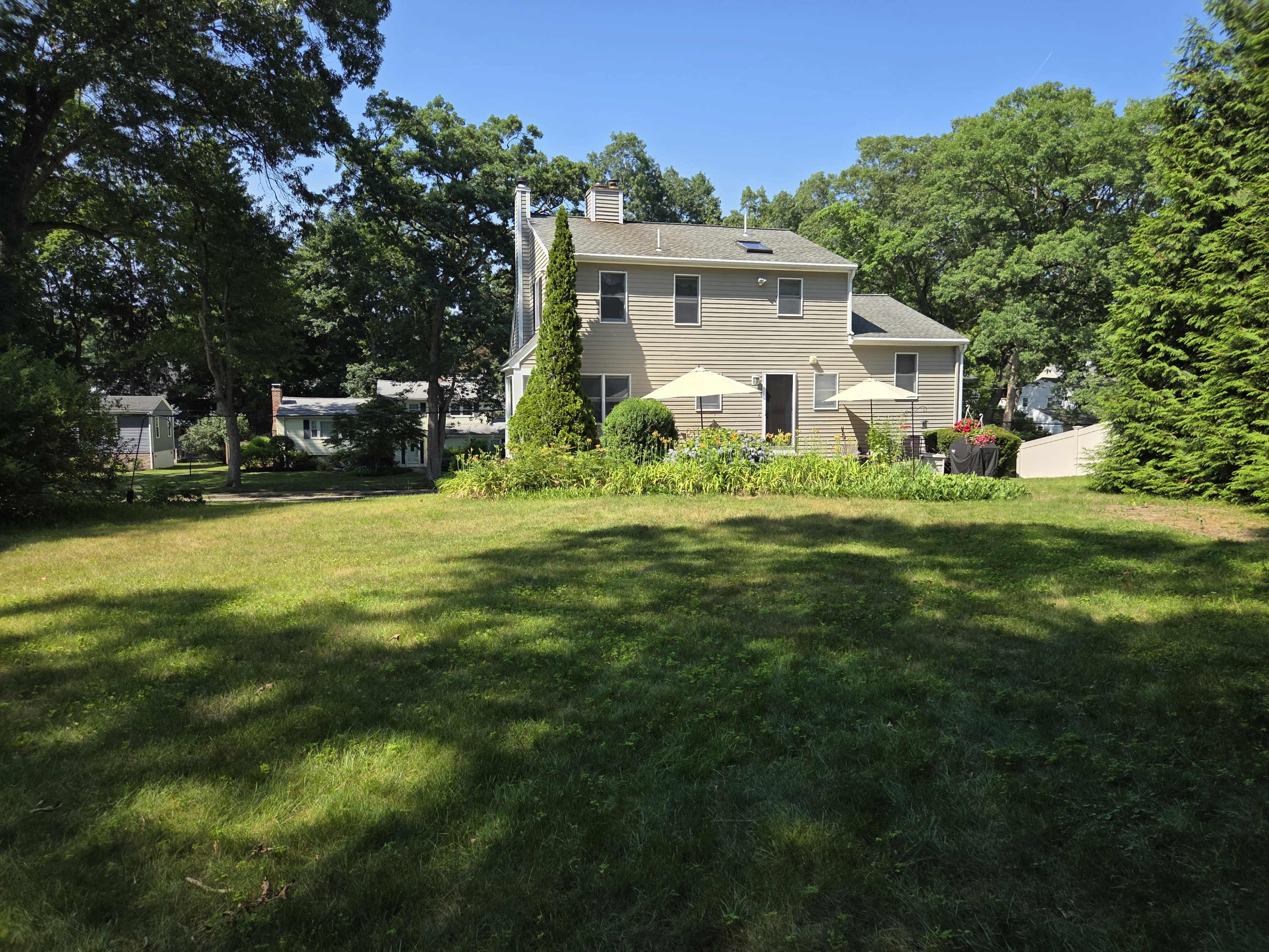 The image shows a spacious backyard with a two-story yellow house and several trees, along with a landscaped area featuring flowers and patio umbrellas.