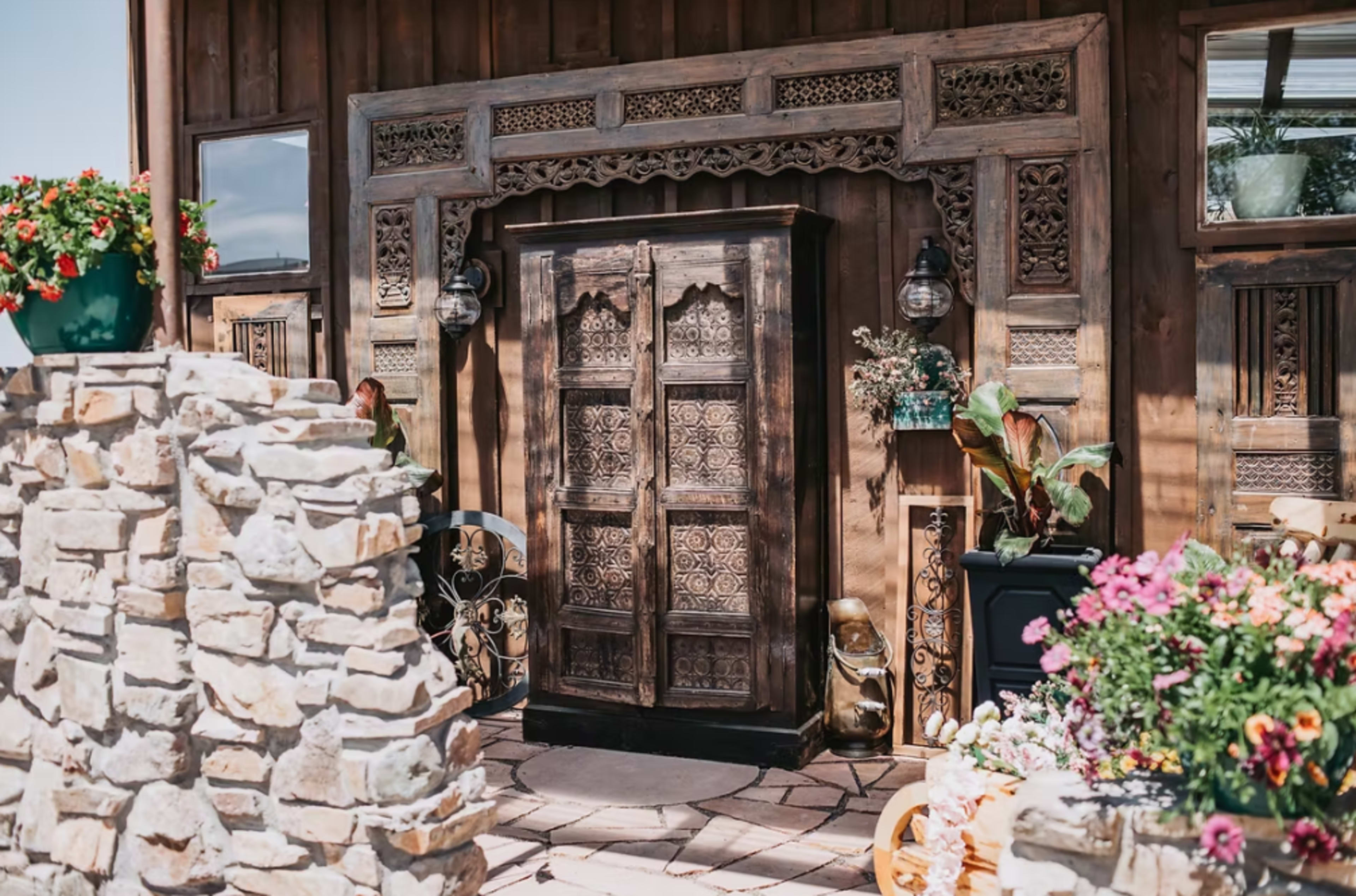 A wooden cabinet with intricate carvings stands against a backdrop of stone and wooden architecture, surrounded by potted plants and flowers.