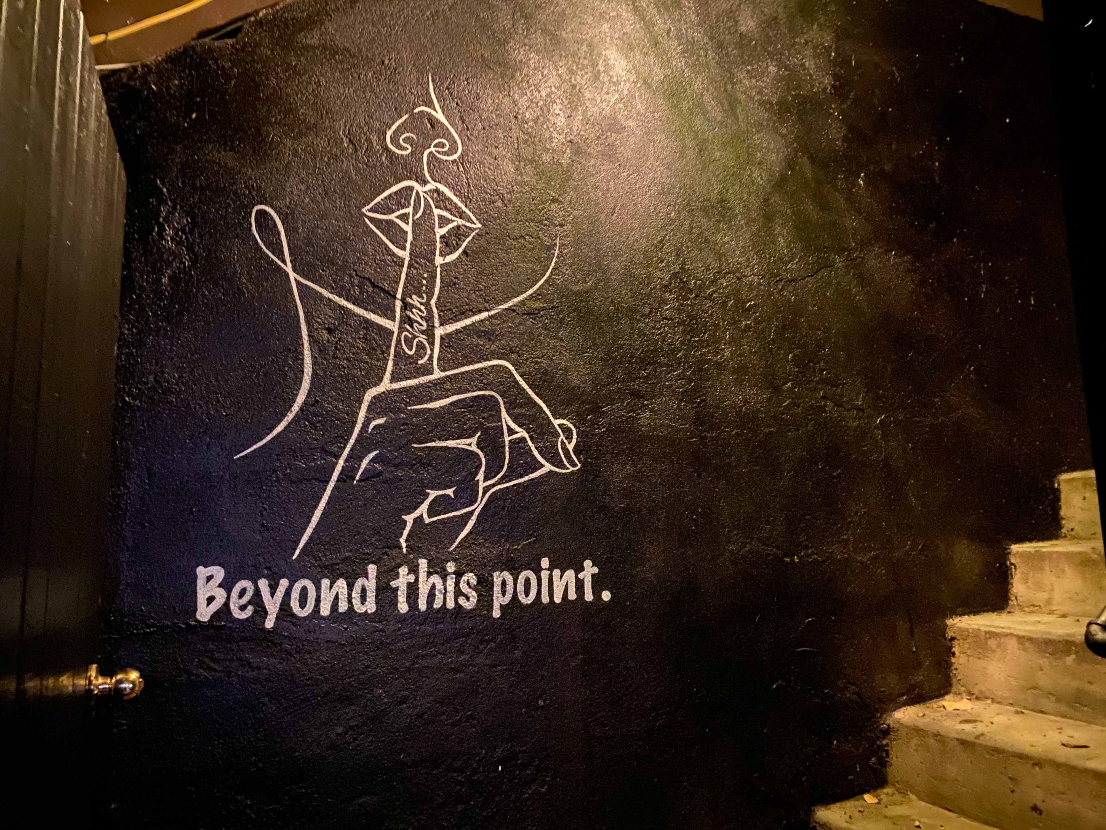 A mural featuring a hand with a finger pressed to the lips is painted on a black wall, accompanied by the words "Beyond this point."