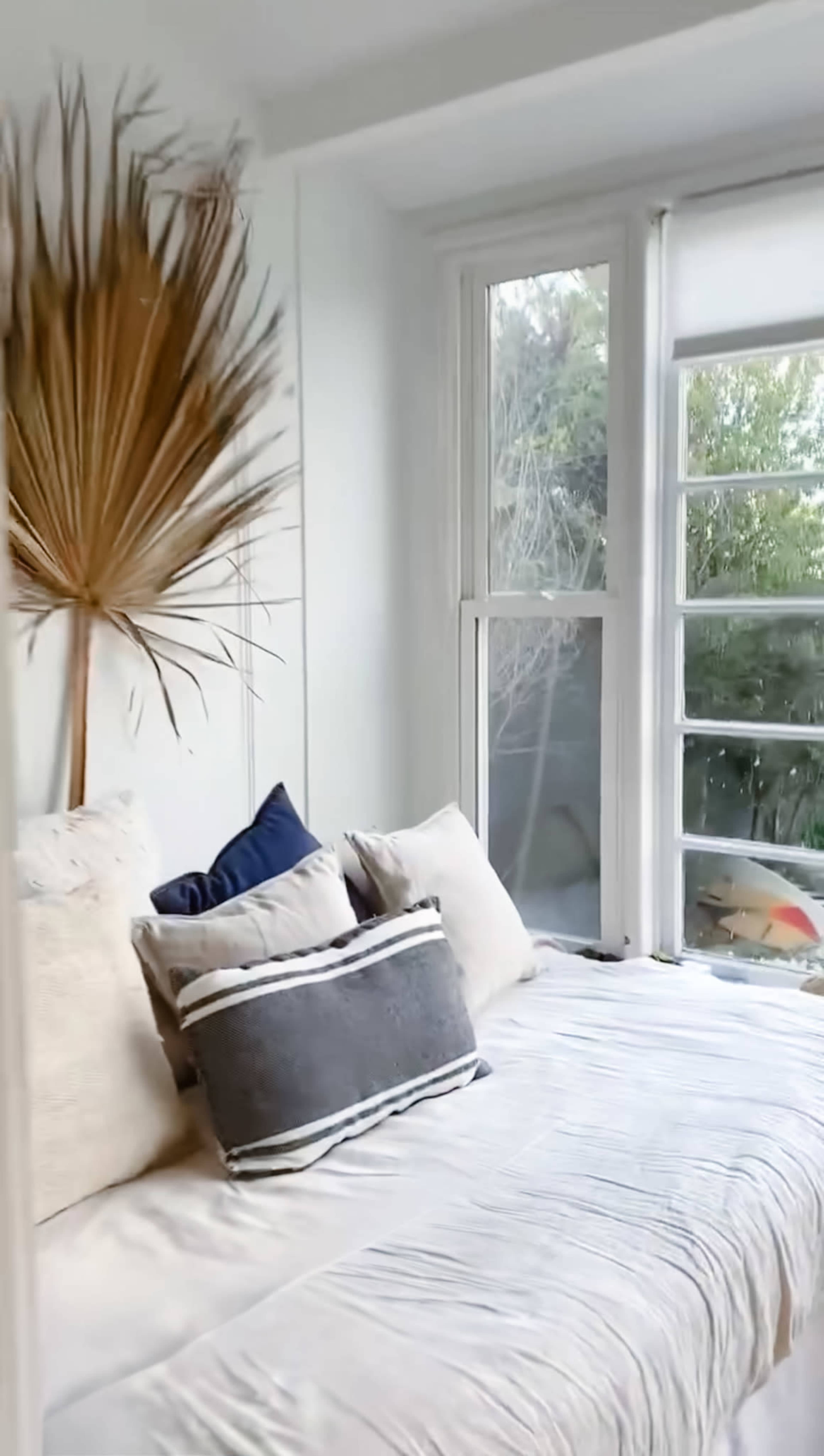 The image shows a cozy bedroom with a large window, a neatly made bed adorned with multiple pillows, and a decorative dried plant in the corner.