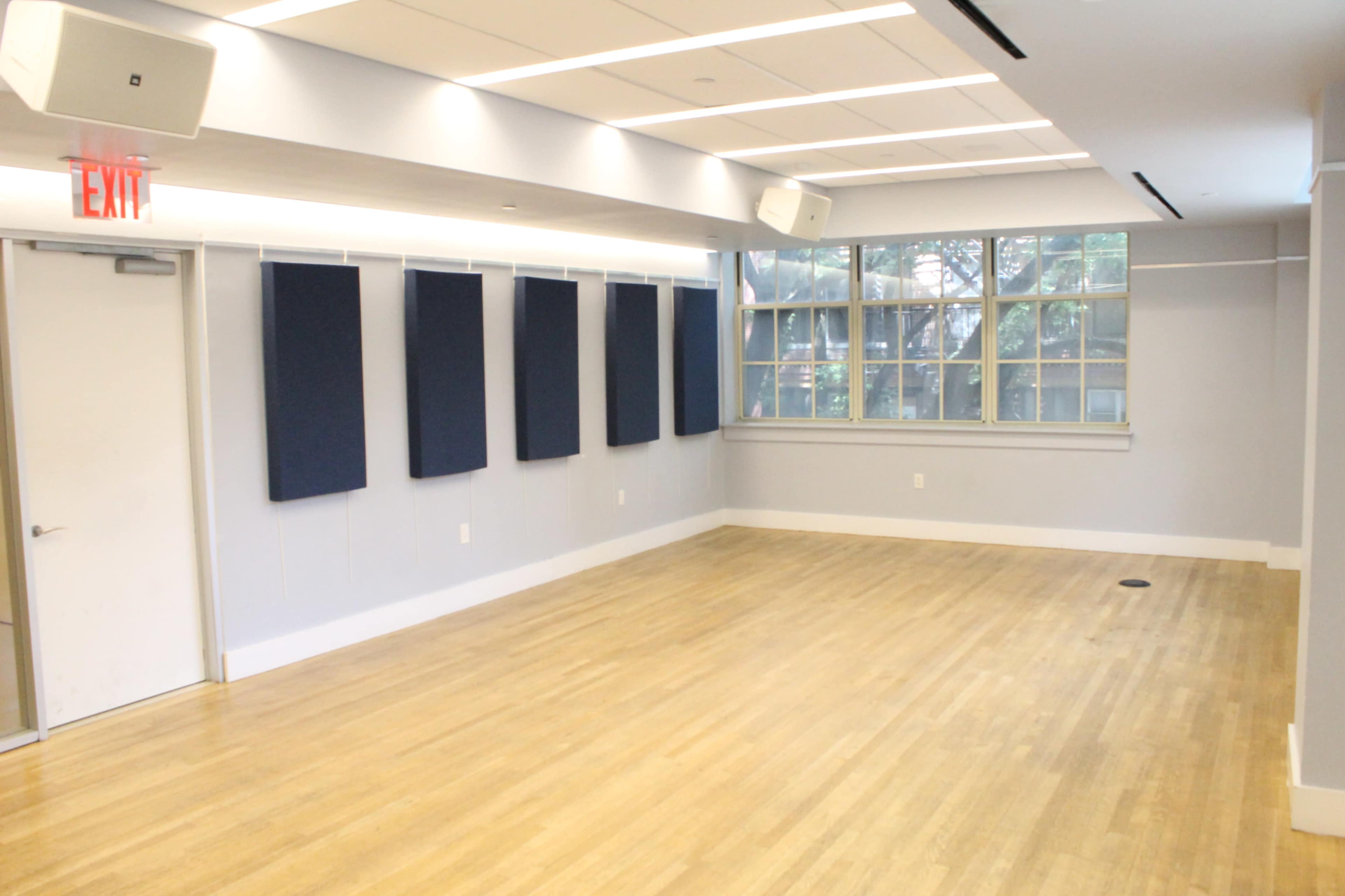 The image shows a spacious, empty room with wooden flooring, white walls, and large windows, featuring several blue sound panels hanging on the wall.