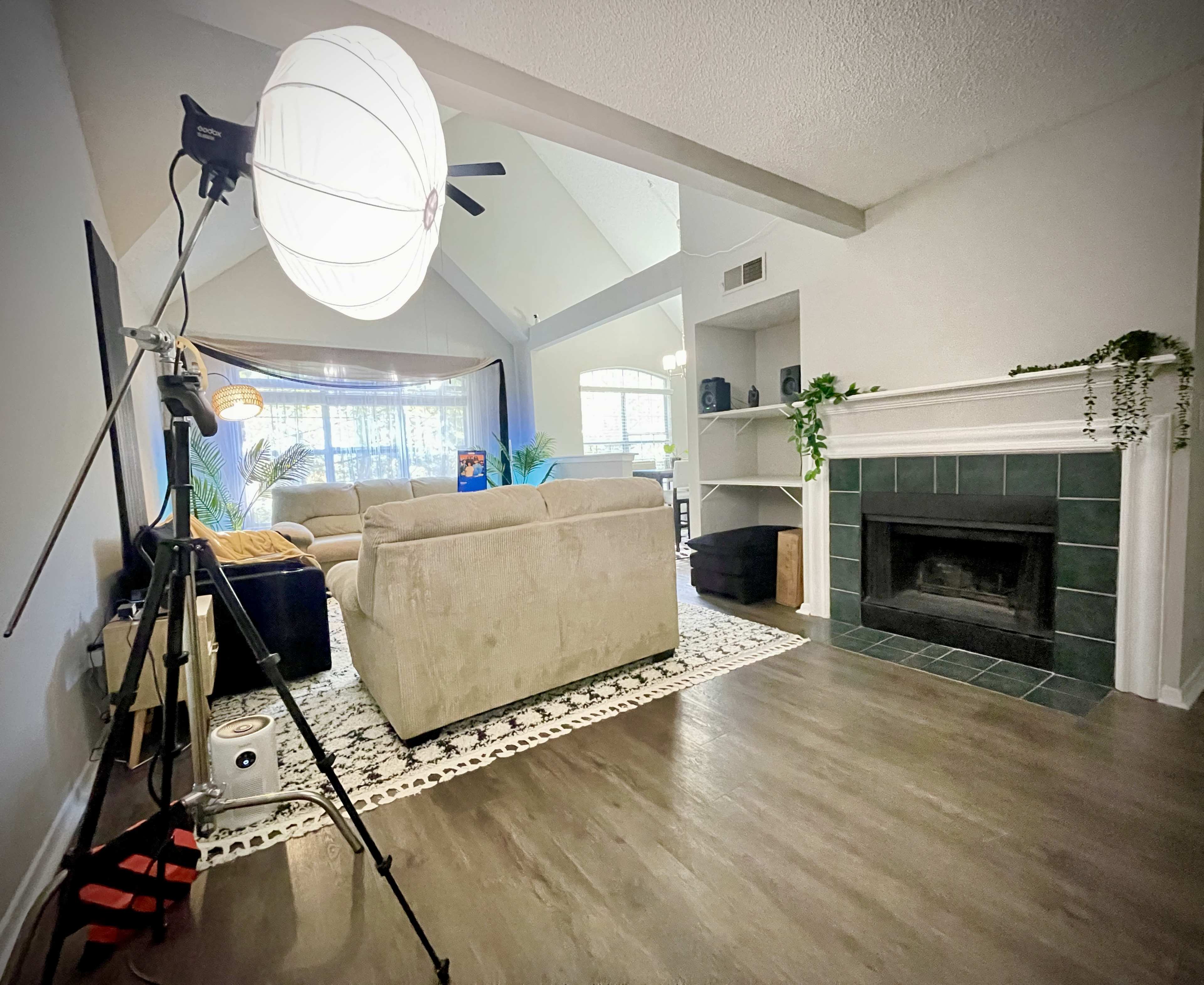 A cozy living room features a large couch, a decorative rug, and a fireplace, with a camera setup and lighting on the left.