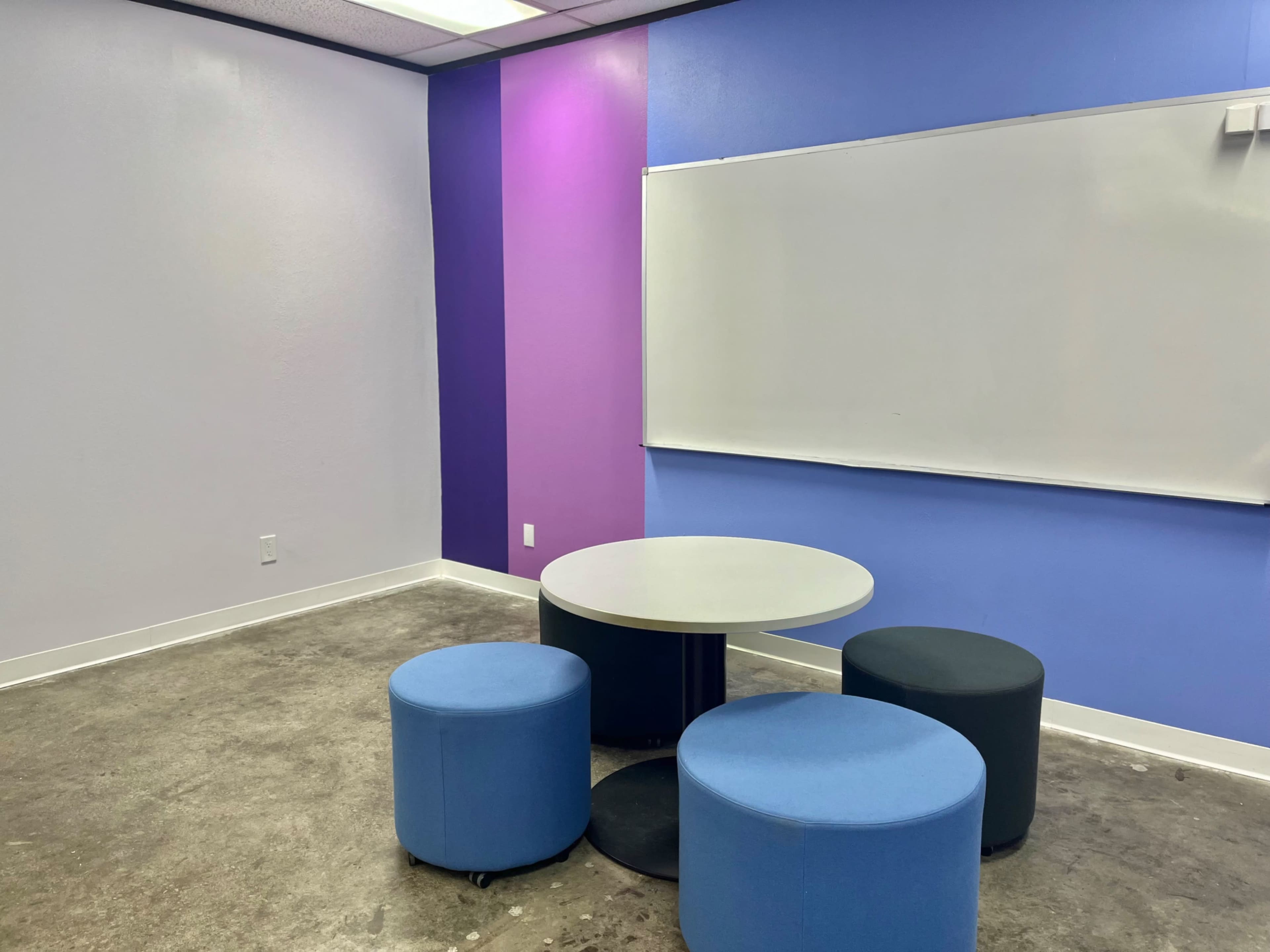 The image shows a small room with a round table and four cylindrical stools arranged around it, featuring colorful walls in shades of purple and blue.