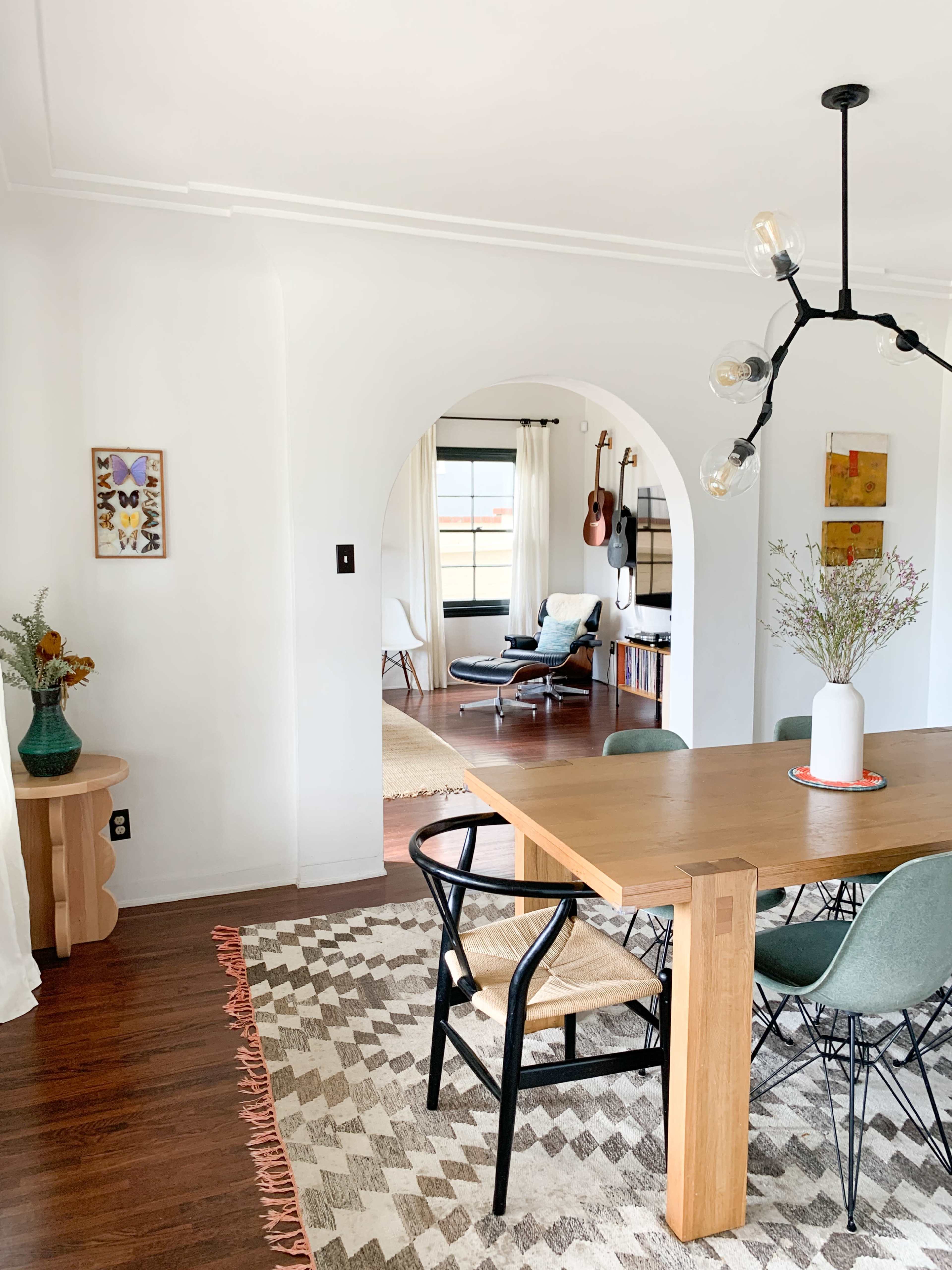 A dining area features a wooden table surrounded by chairs, with an archway leading to a living space that includes guitars and artwork on the walls.