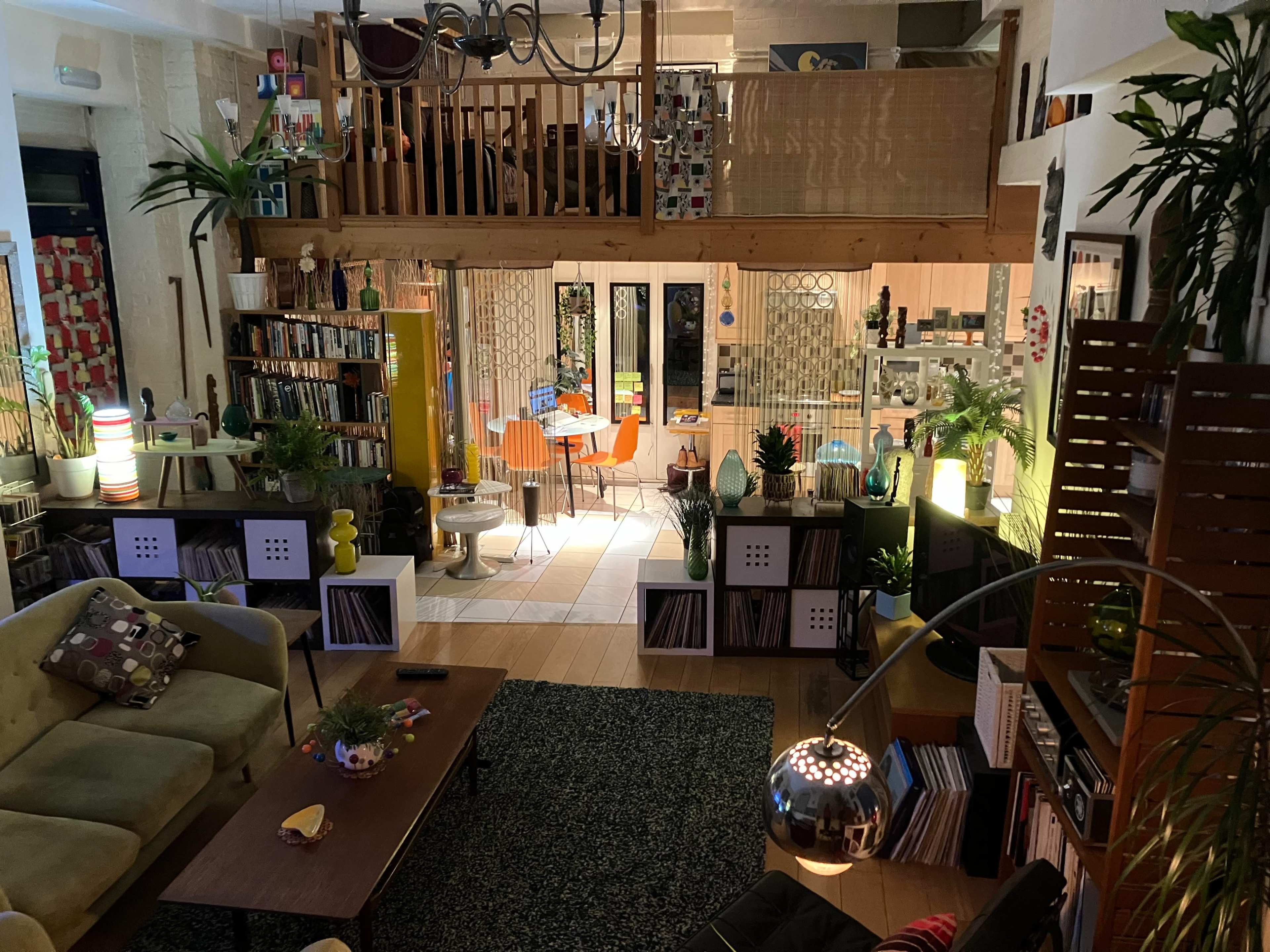 The image shows a spacious, multi-level living area filled with bookshelves, plants, and comfortable seating, with a dining area visible in the background.