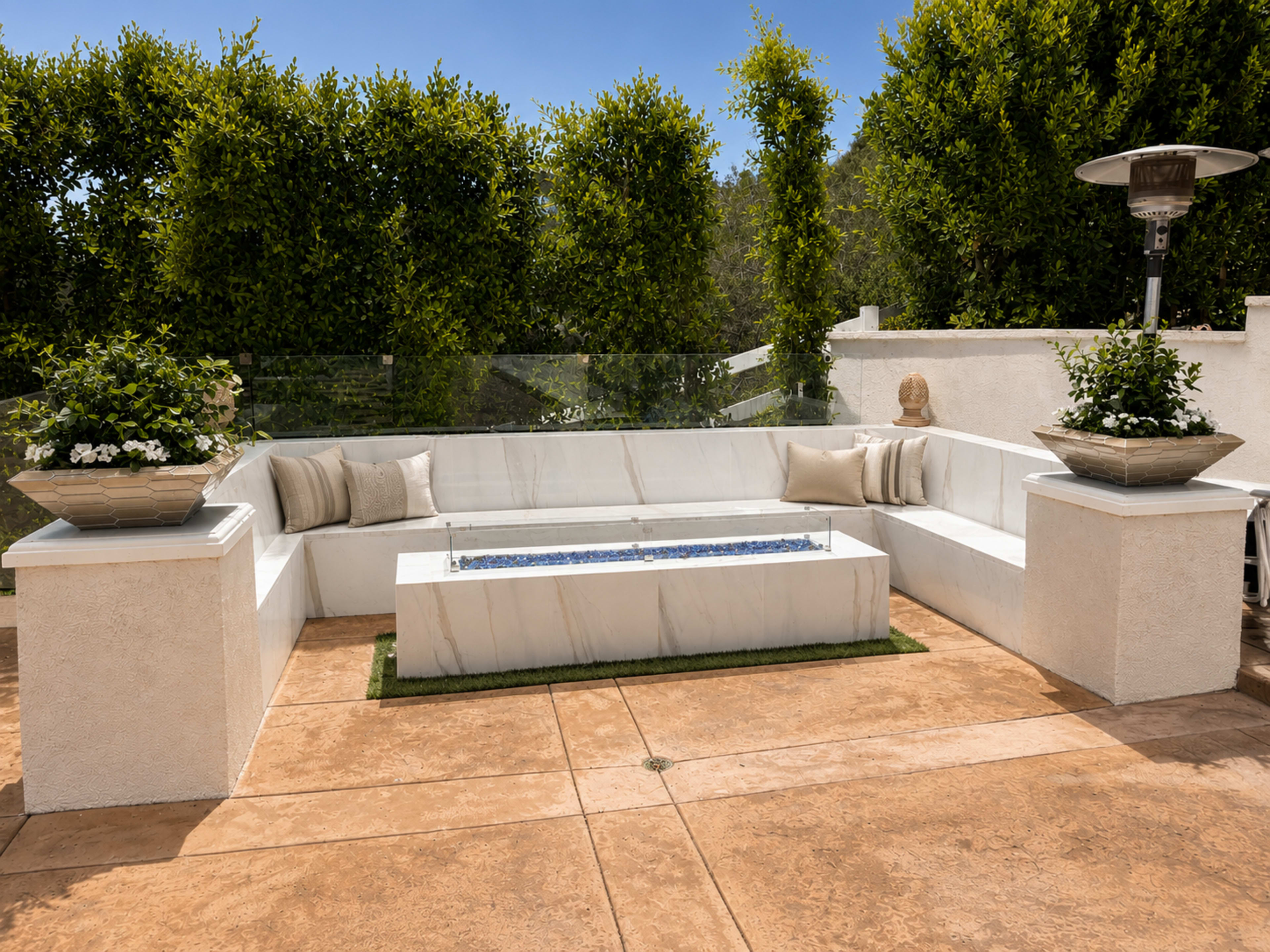 The image shows a modern outdoor seating area with a marble-topped fire pit and surrounding white sectional sofa adorned with planters and cushions.
