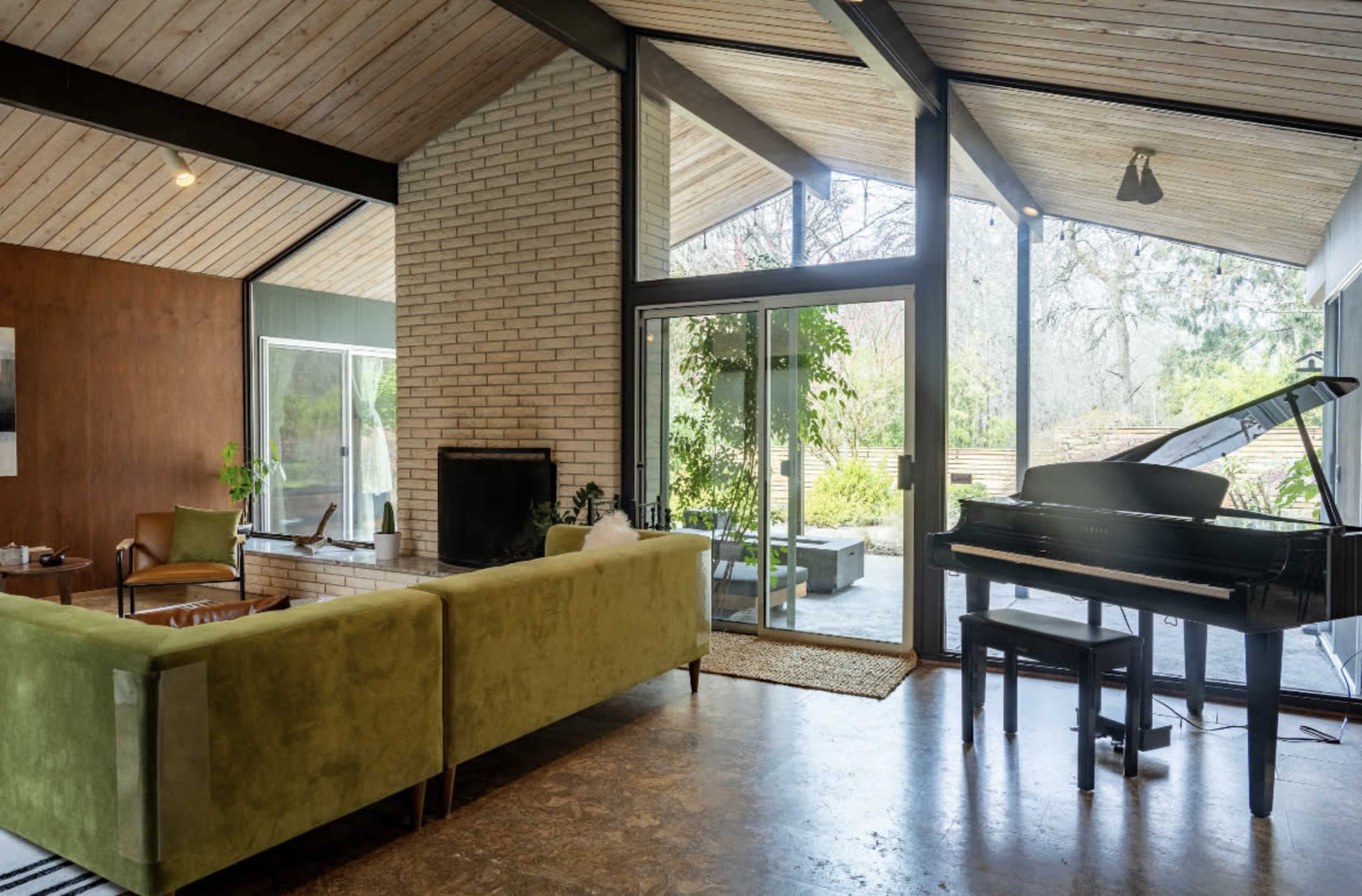 A spacious living room features a green sofa, a black piano, and large glass doors that open to a landscaped outdoor area.