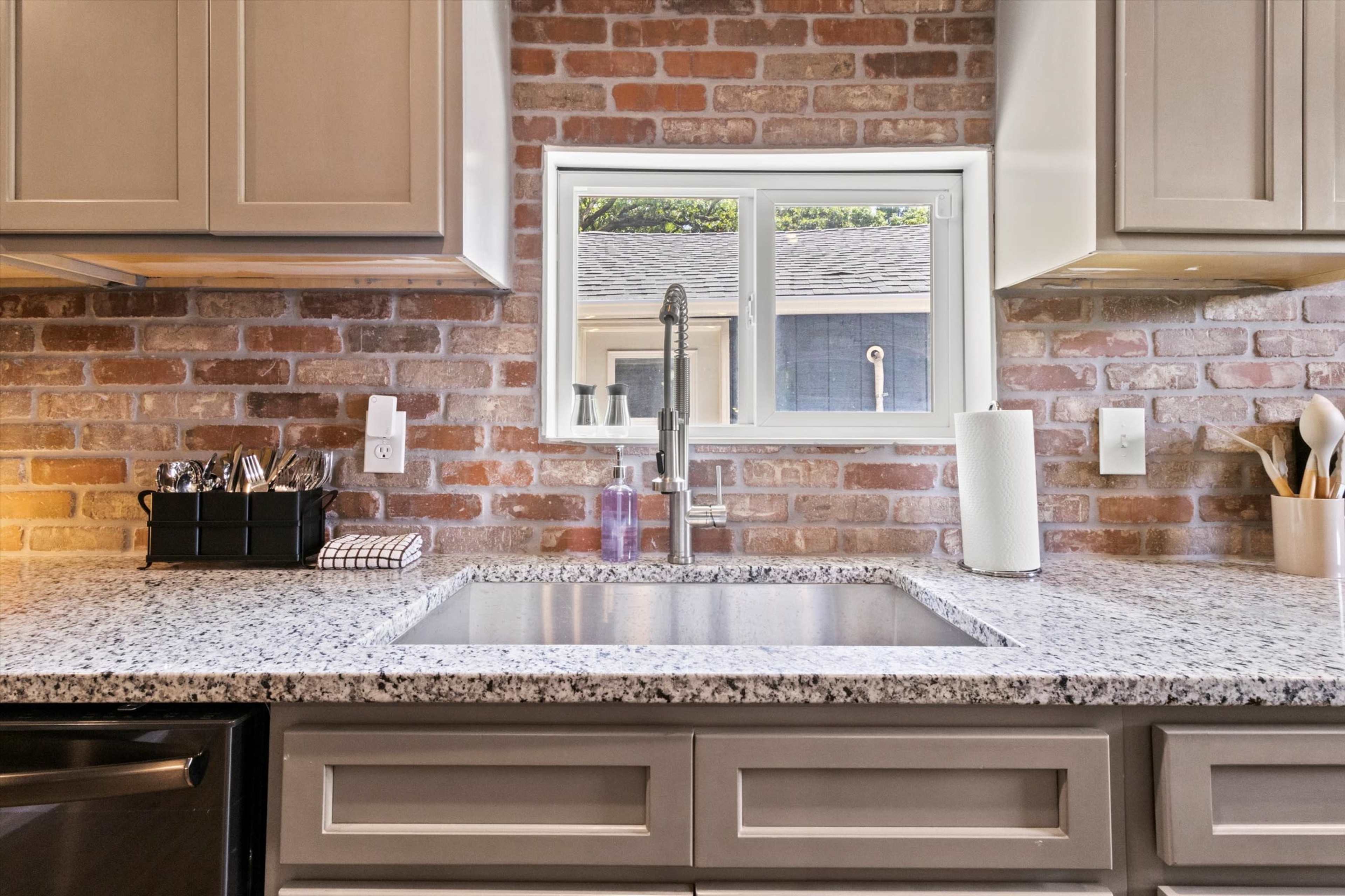 A kitchen features a granite countertop with a central sink, accompanied by a window overlooking a brick wall and stainless steel appliances.