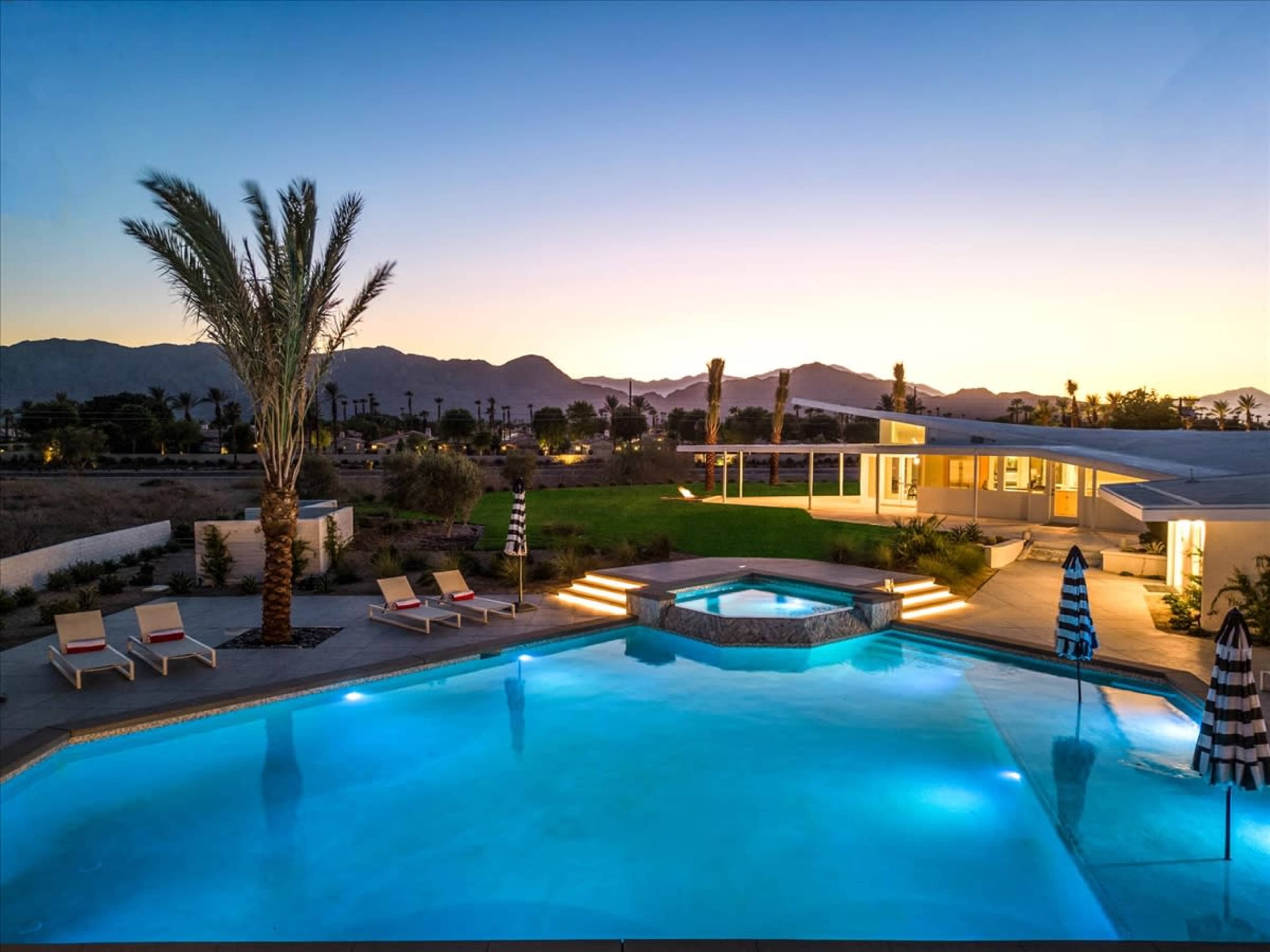 A swimming pool and spa are illuminated beside a modern house with palm trees and mountains in the background during sunset.