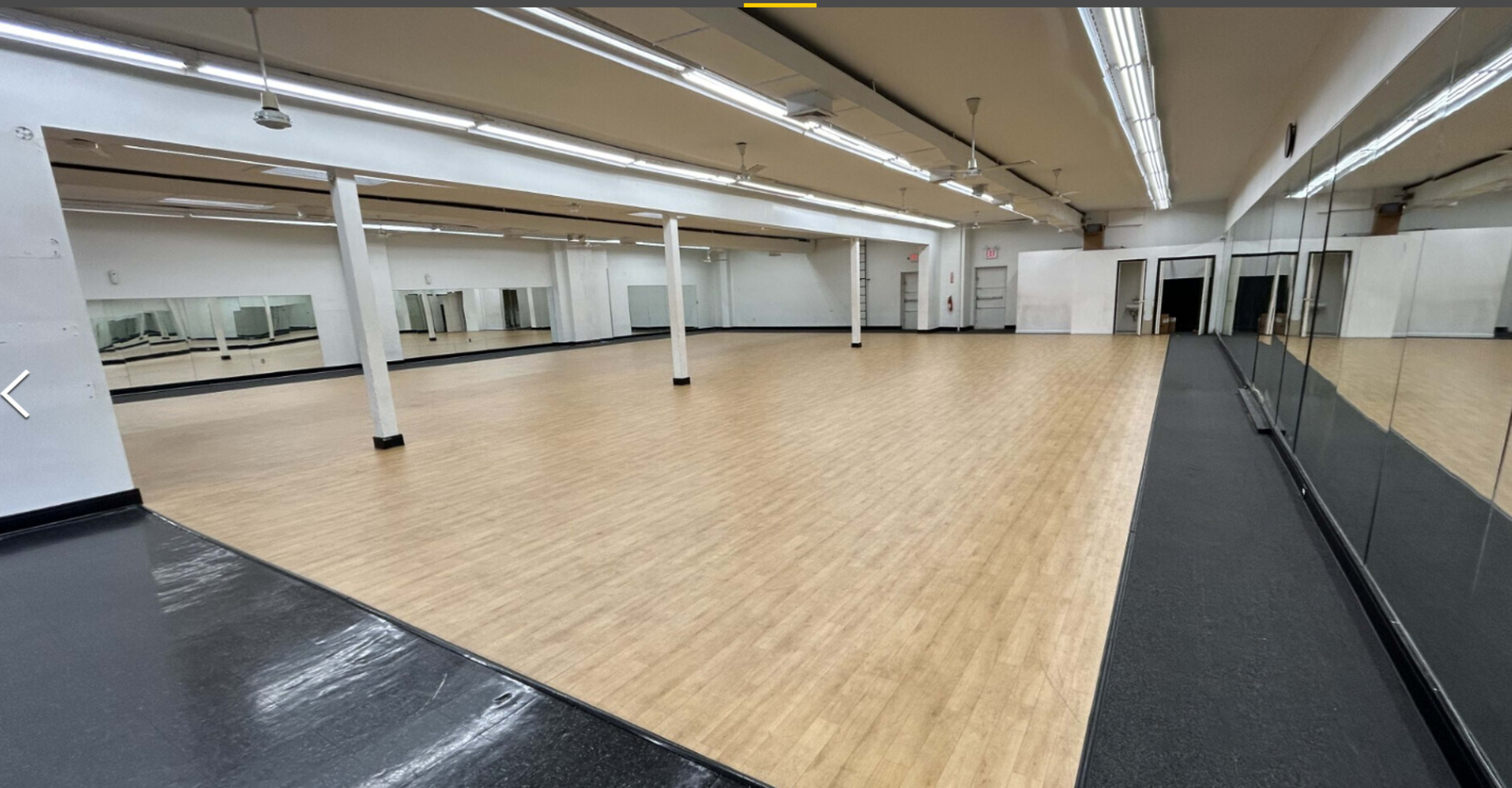 The image shows a large, empty dance studio with wooden flooring, mirrors on one wall, and white walls throughout.