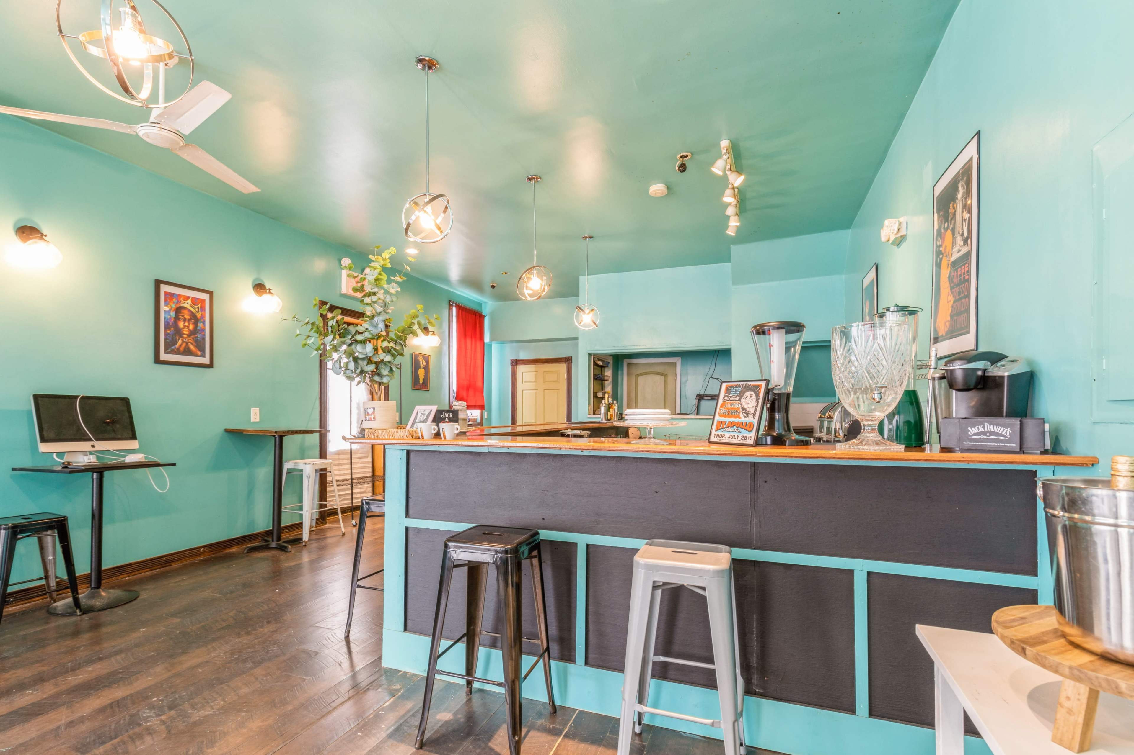 The image shows a brightly colored café with a turquoise interior, featuring a wooden counter, black and silver stools, and modern lighting fixtures.