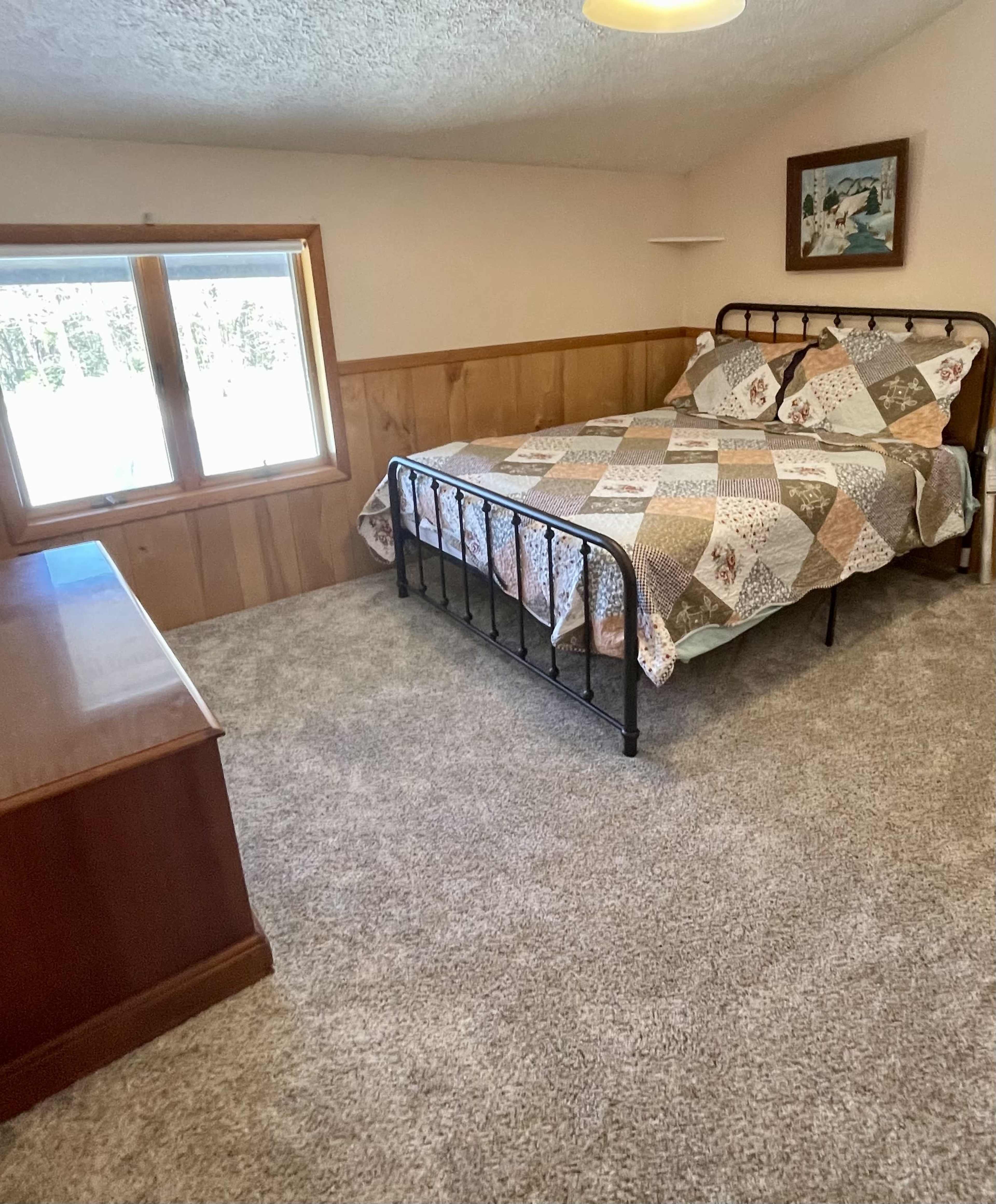 A bedroom features a metal bed with a patchwork quilt, a dresser, and a window overlooking a wooded area.
