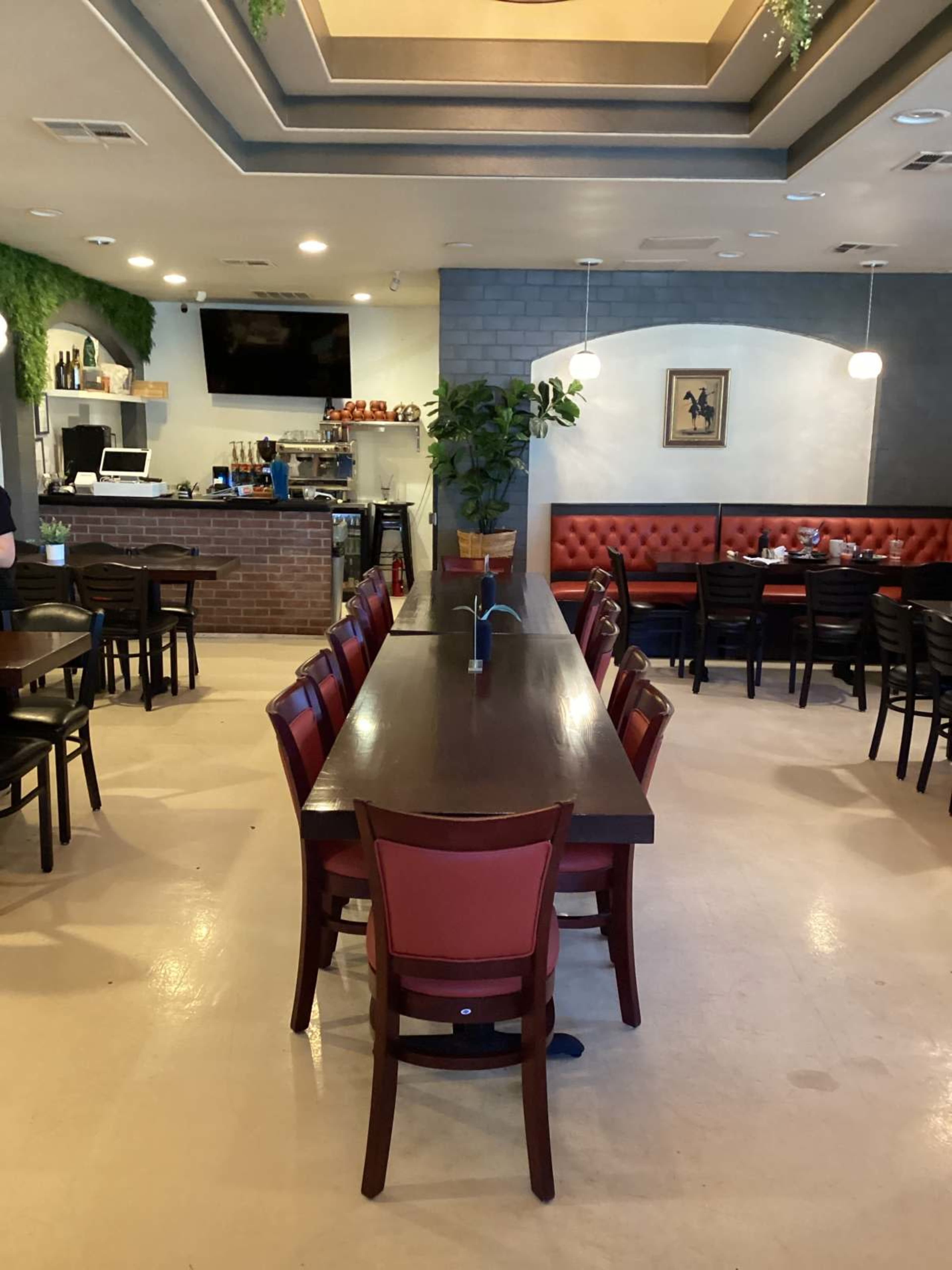 The image shows a restaurant interior featuring a long communal table surrounded by chairs, with a bar area in the background and booths along the side.