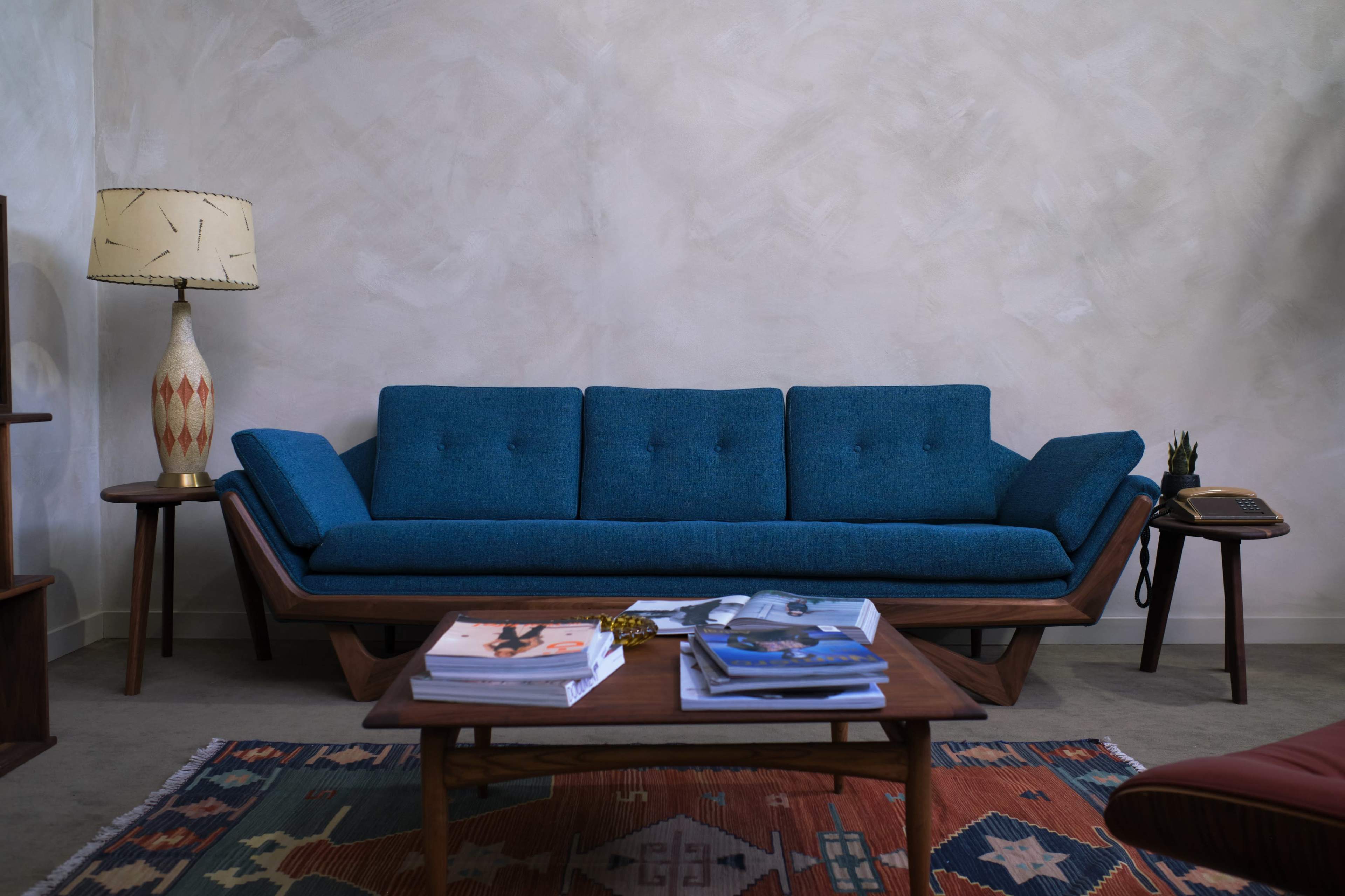 The scene features a blue mid-century modern sofa, a patterned area rug, a wooden coffee table with magazines, and a standing lamp against a textured light wall.