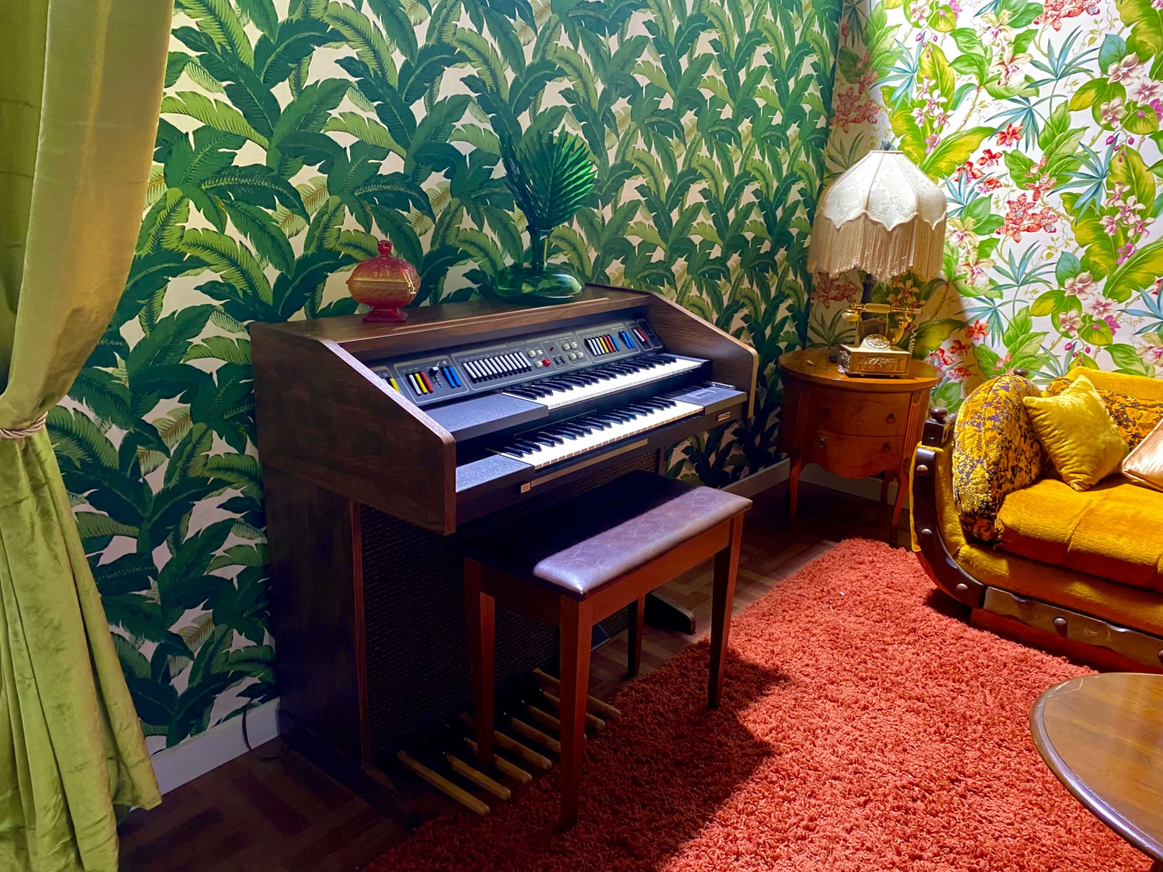 A vintage organ sits on a wooden stand in a room with leafy green wallpaper and a retro orange sofa.