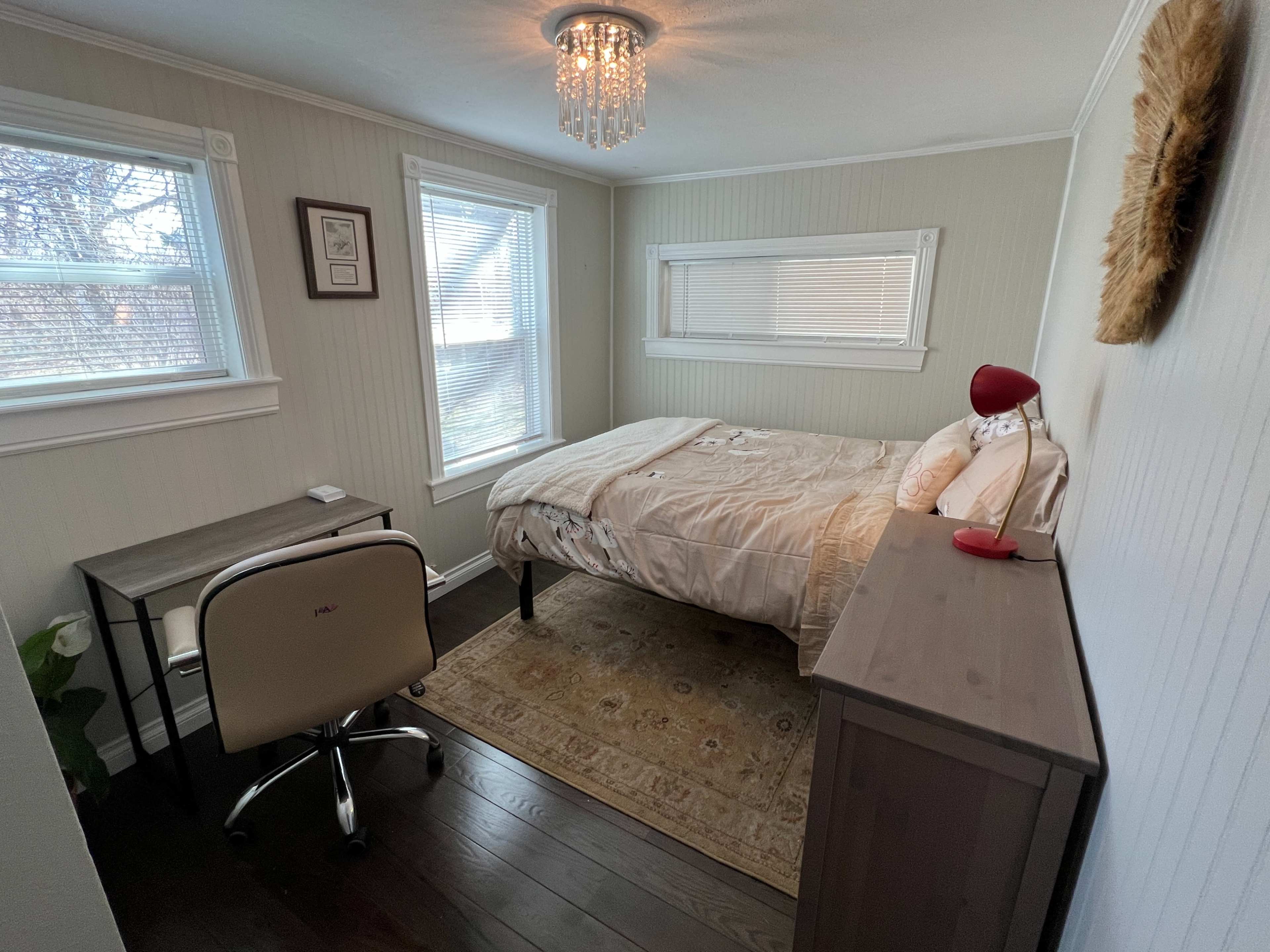 A small, well-lit bedroom featuring a bed, a desk with a chair, and a wooden dresser.