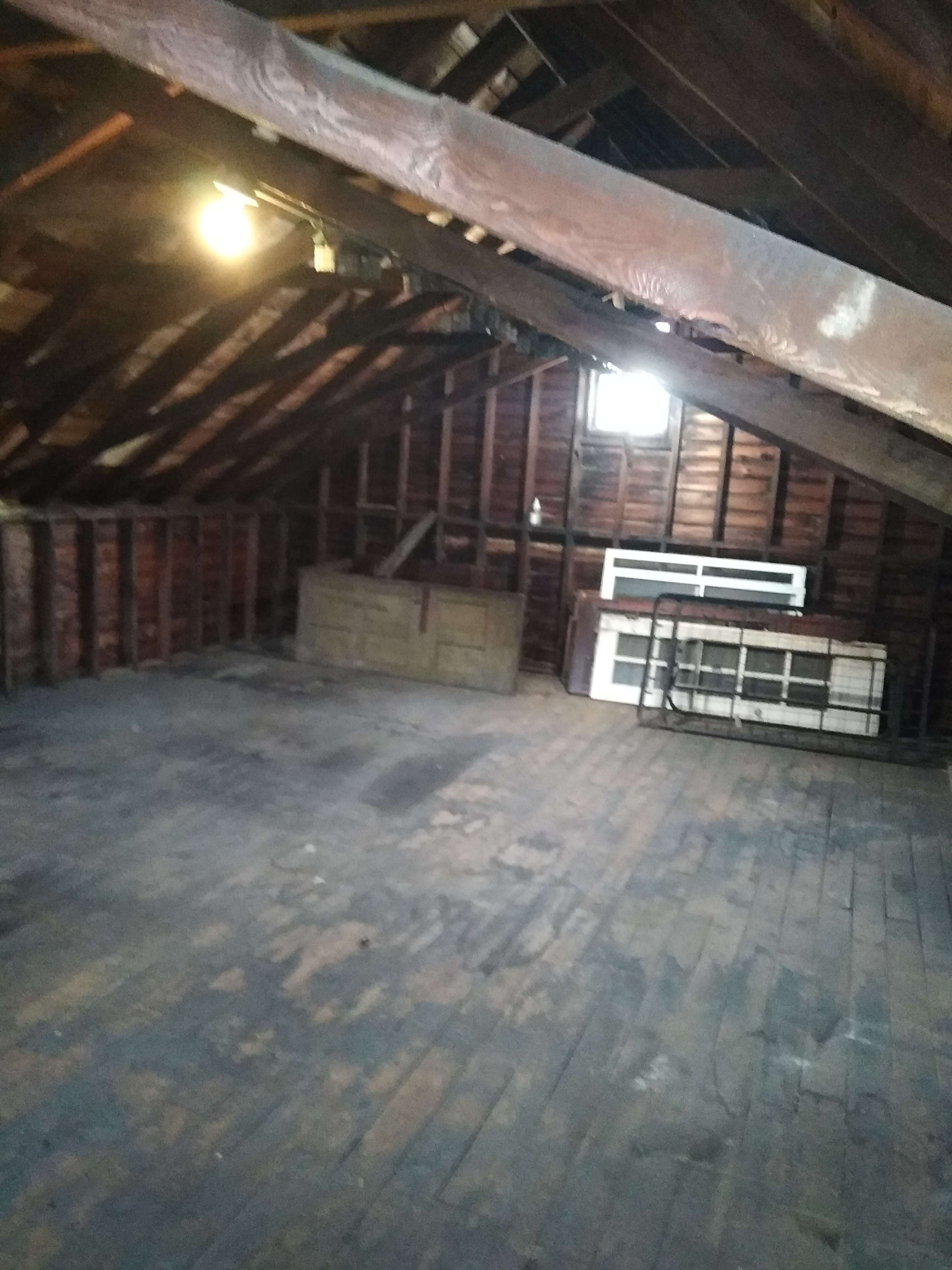 A spacious, unfinished attic with exposed wooden beams and a wooden floor, containing a storage chest and a white frame against the wall.