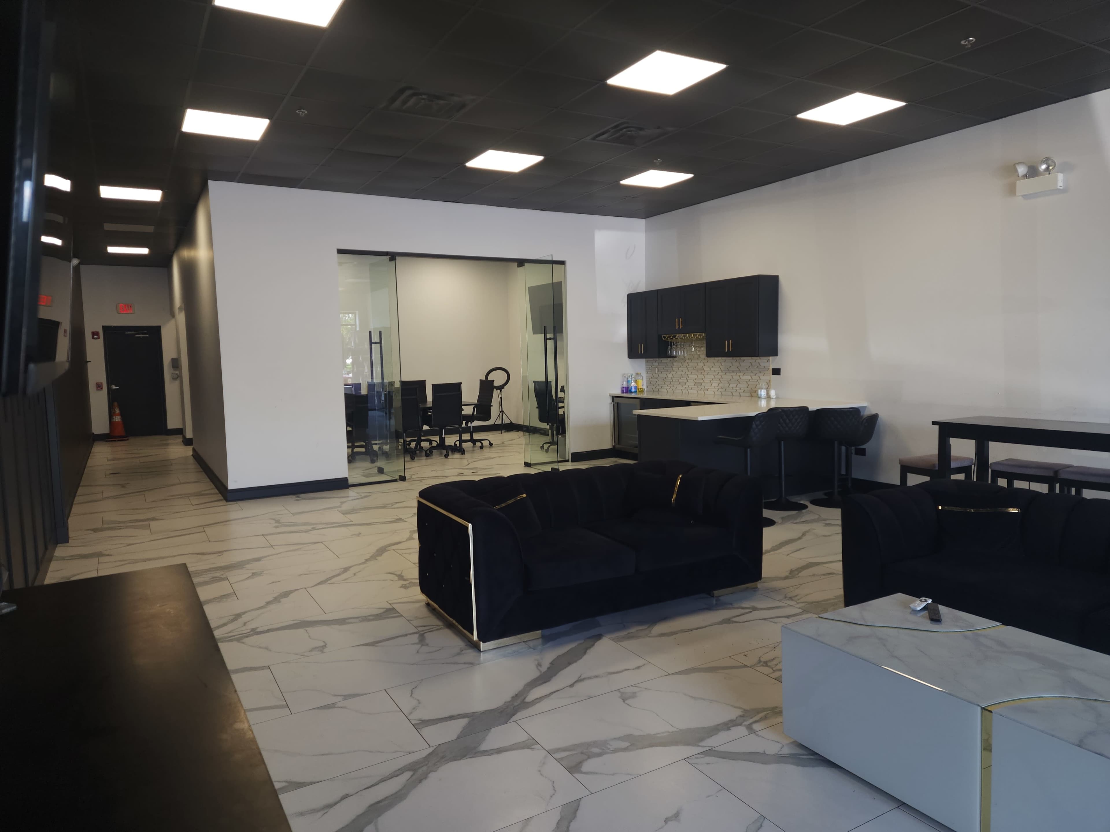The image shows a modern office lounge with dark furniture, a kitchenette, and a glass-walled meeting room in the background.