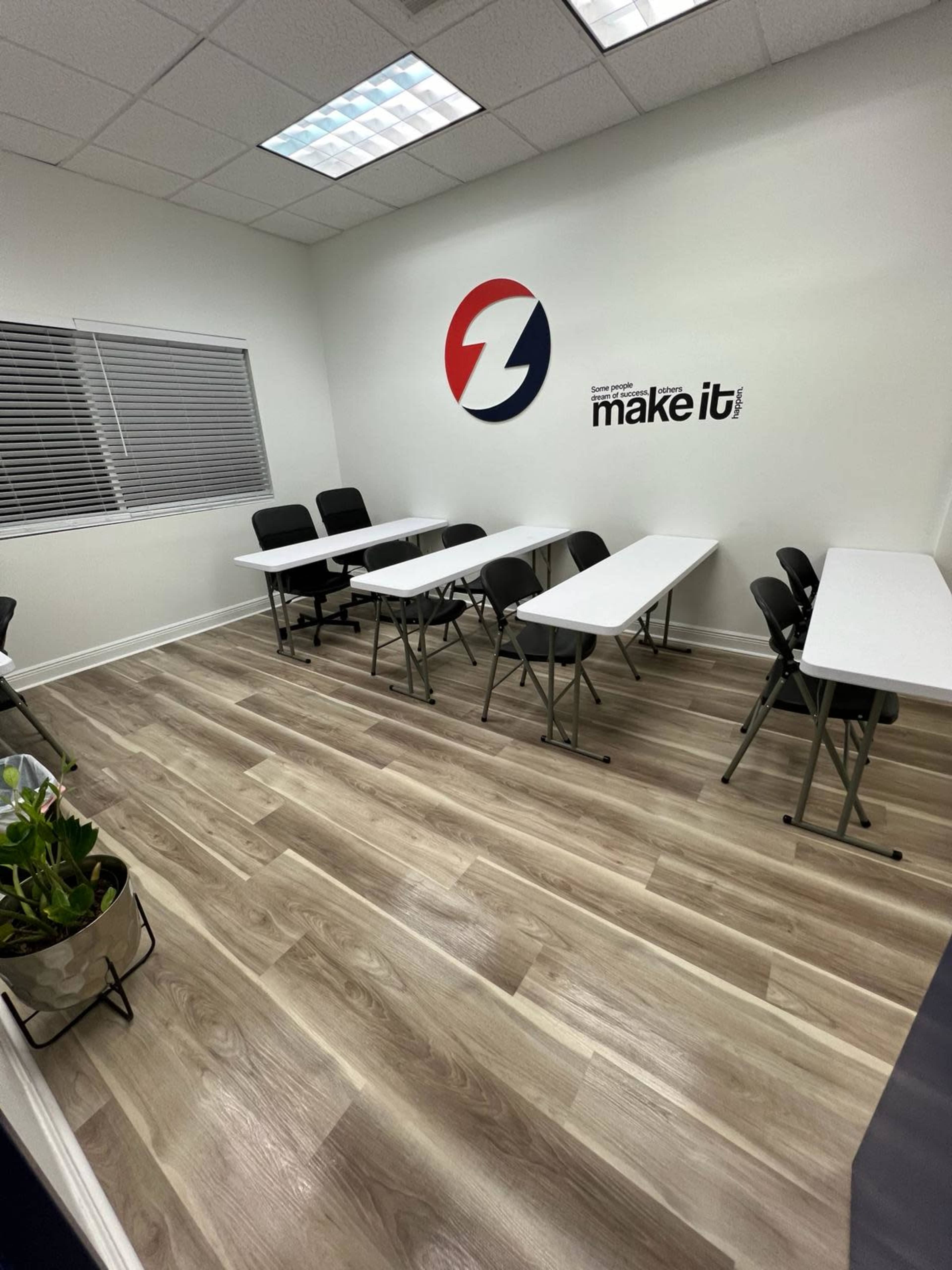 A small meeting room features several folding tables and chairs arranged neatly, with light-colored wooden flooring and a wall displaying a logo and the phrase "make it."