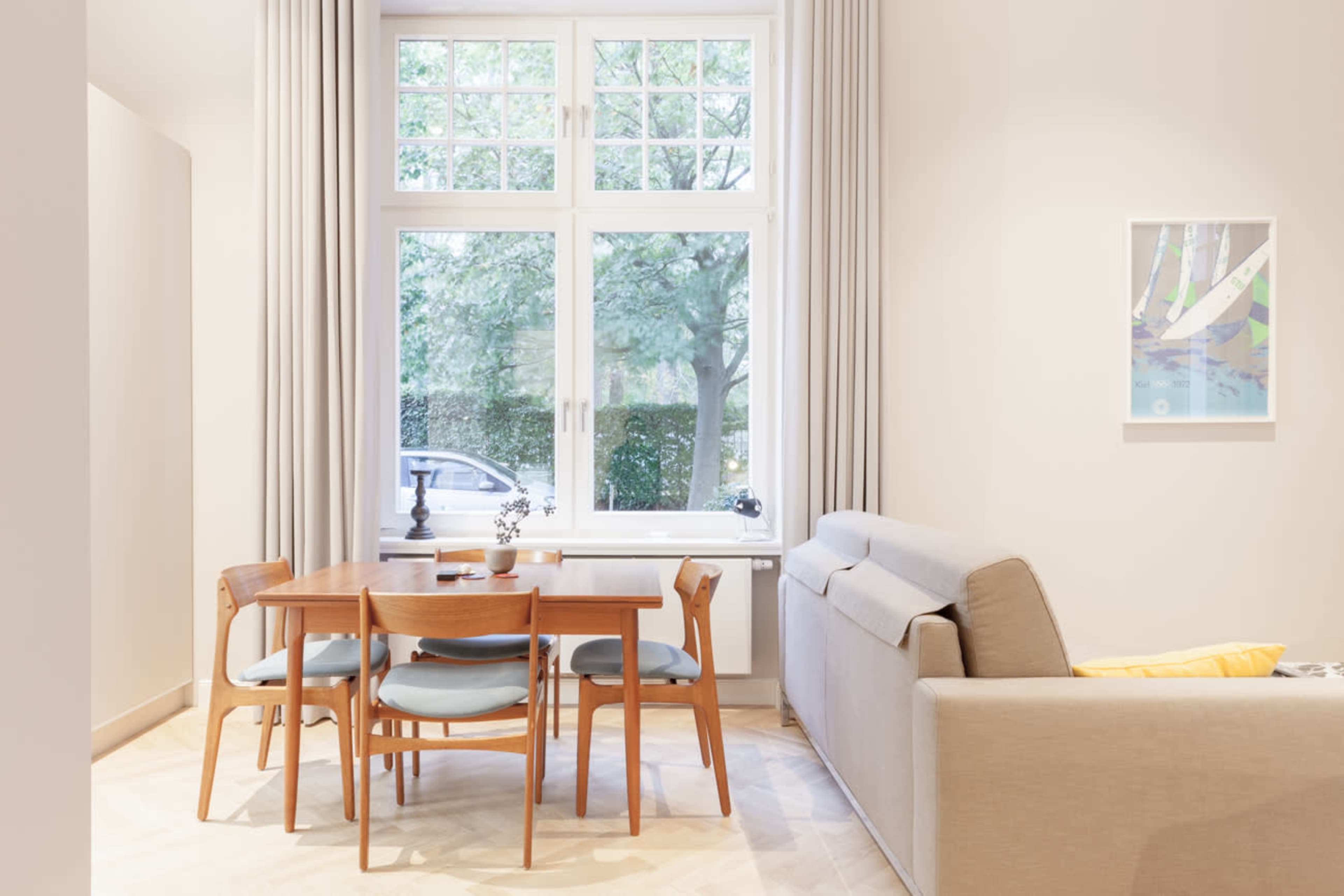 A dining area features a wooden table with four blue chairs beside a large window overlooking a tree-lined street.
