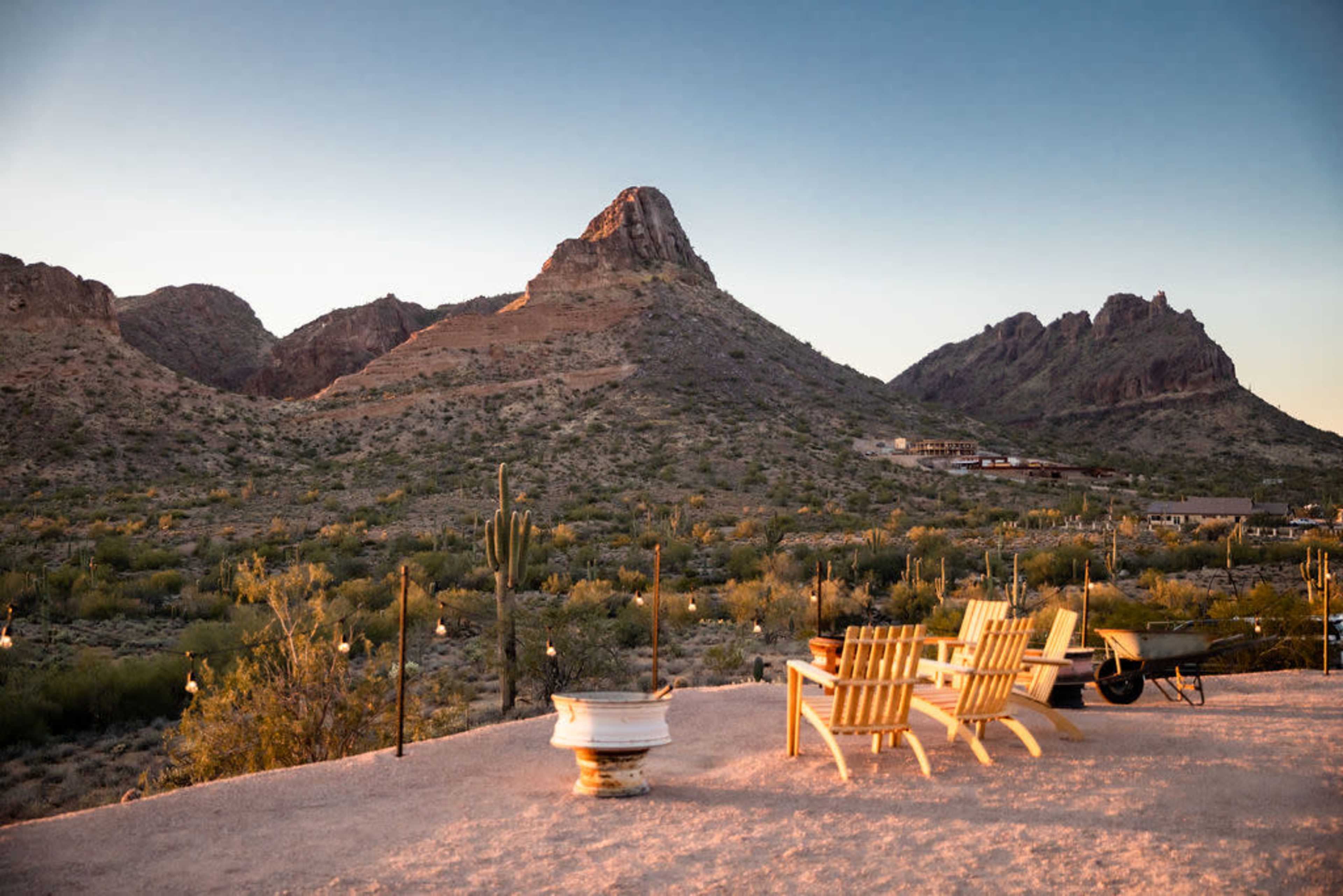 A patio with wooden chairs overlooks a desert landscape featuring mountains and cacti at sunset.