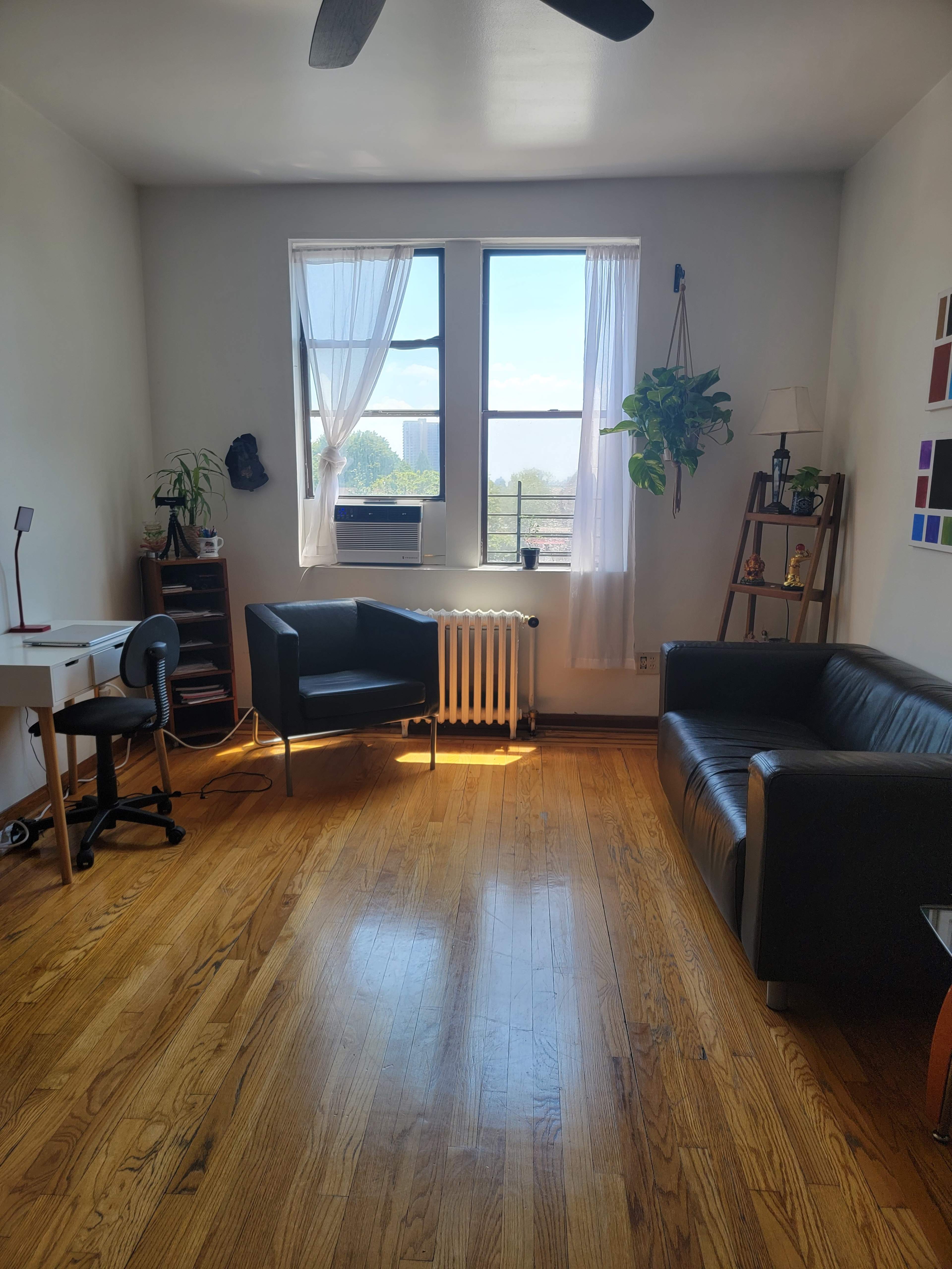 A bright room with hardwood floors, featuring a desk and chair, two seating areas, and large windows with sheer curtains.