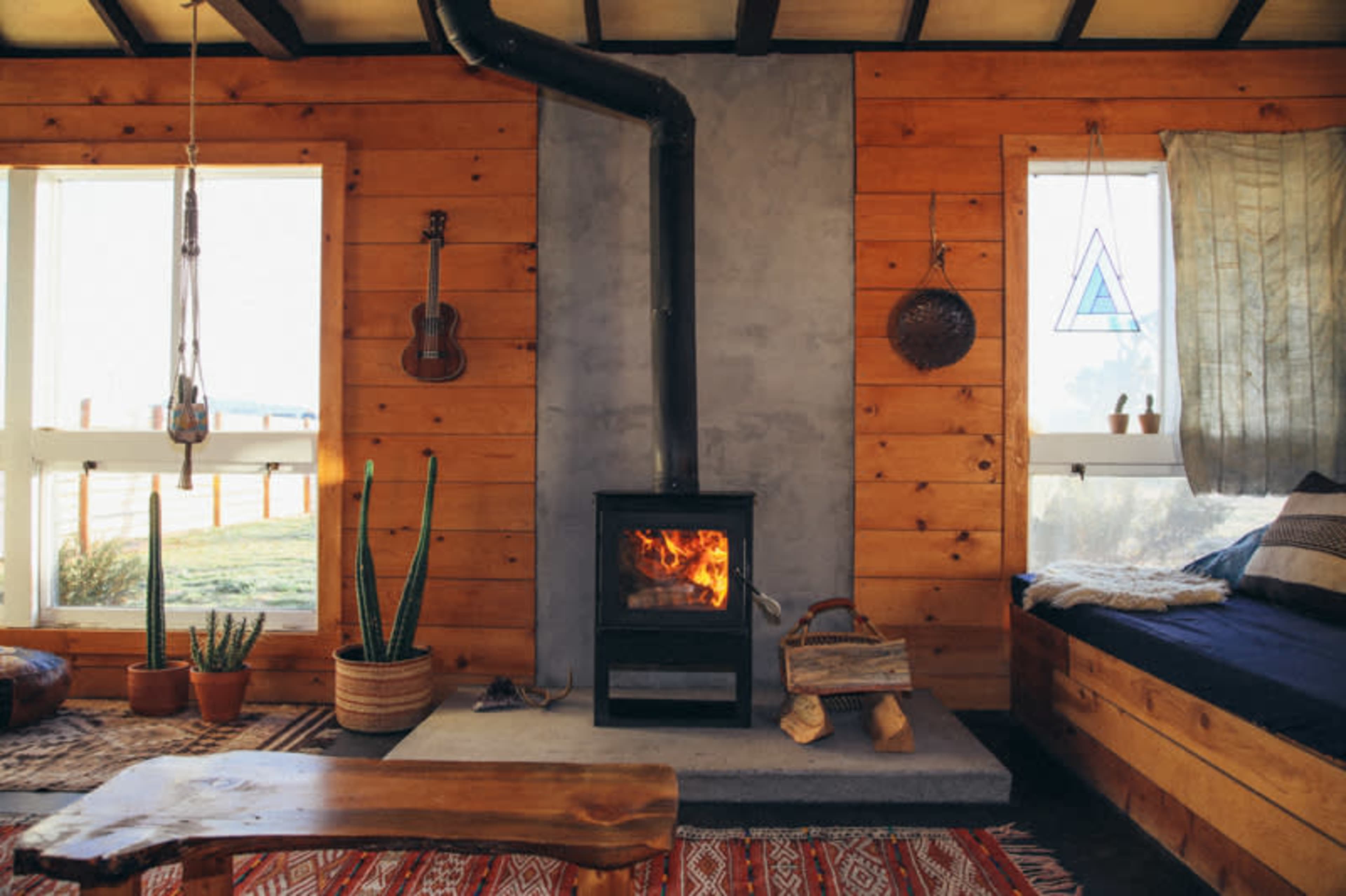 The room features a wood-burning stove as a focal point, surrounded by wooden walls, large windows, and various plants.