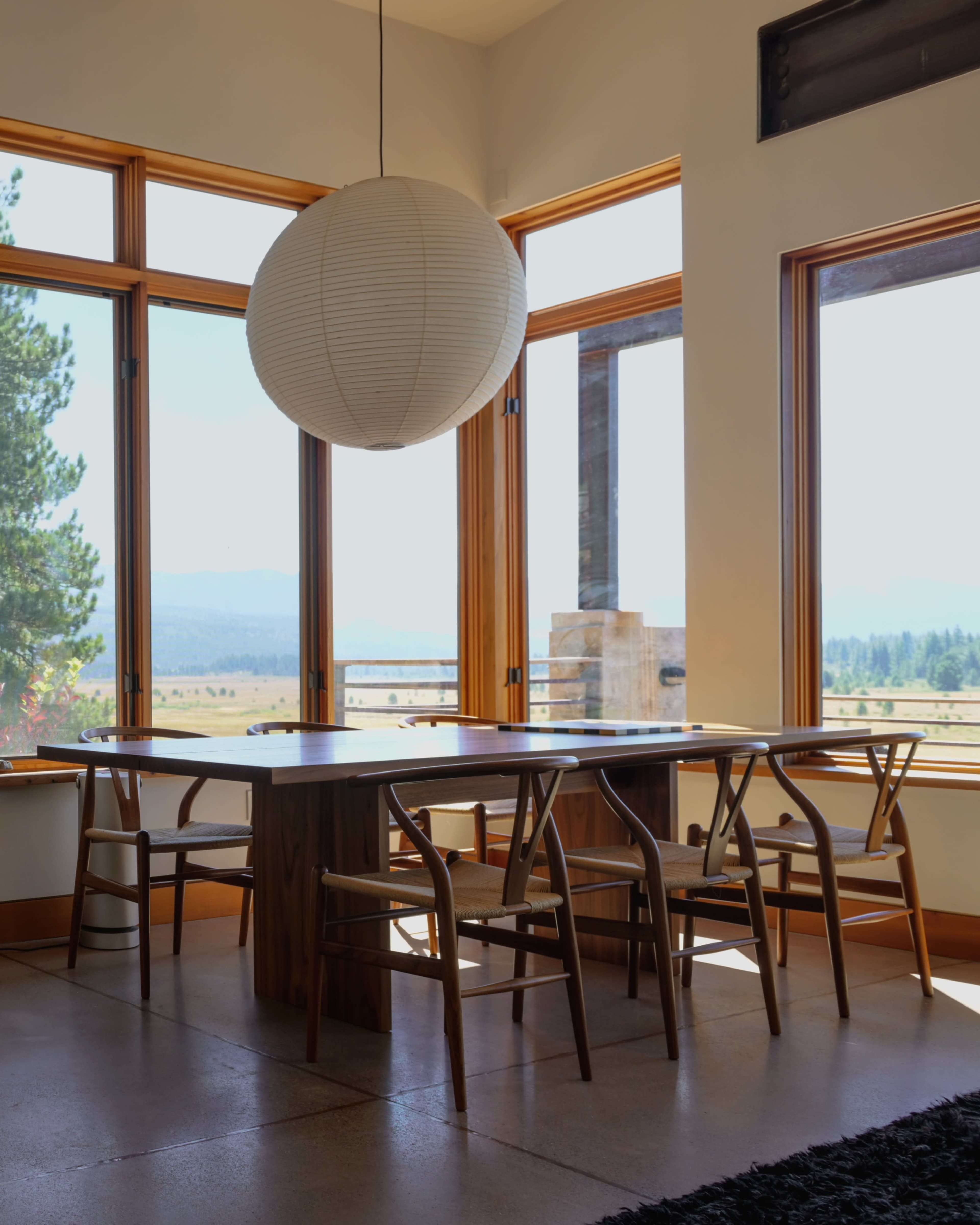 A modern dining area features a wooden table surrounded by chairs and a large pendant light, with large windows offering a view of the landscape.