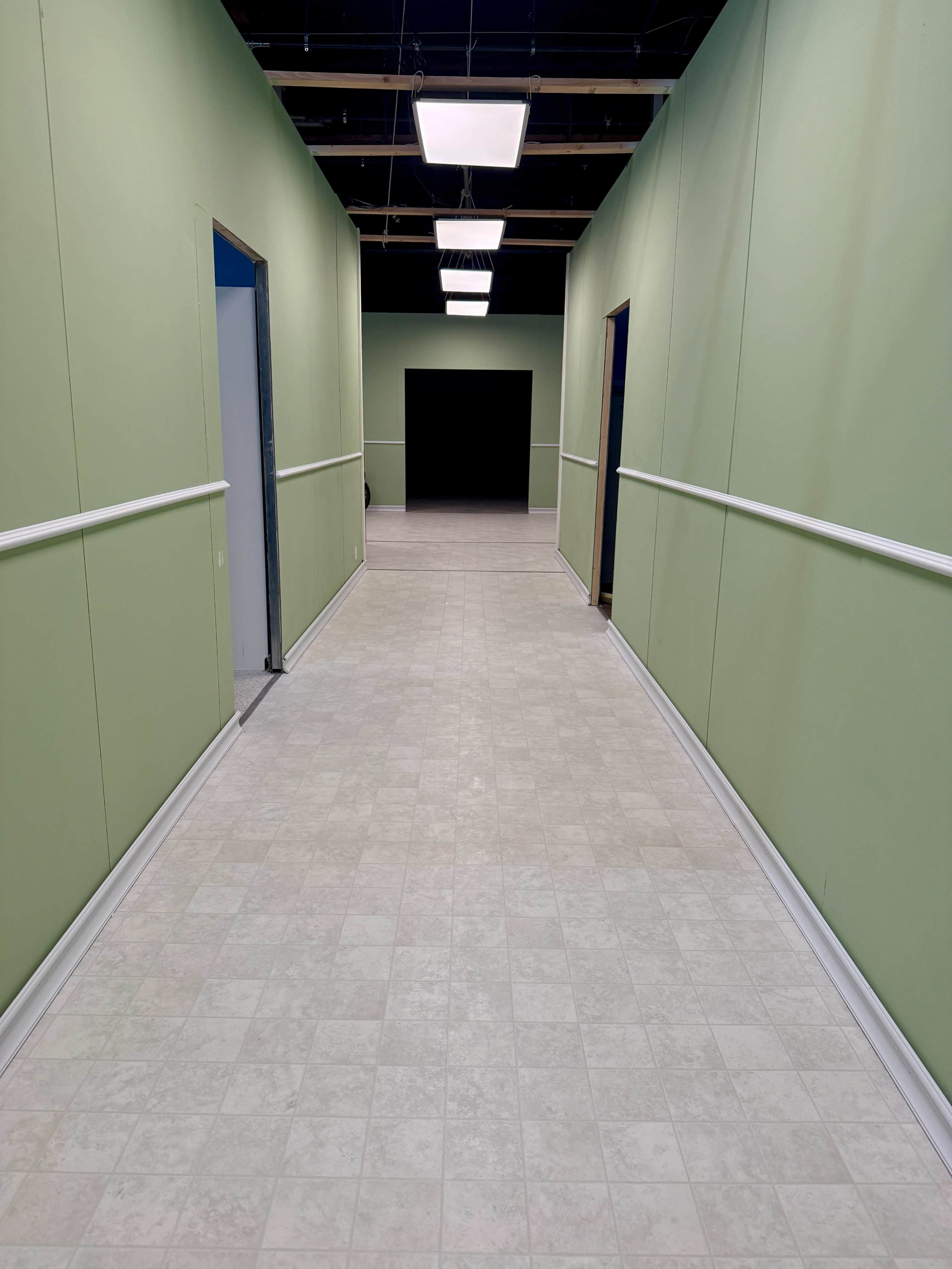 The image shows a long, empty hallway with pale green walls, a tiled floor, and light fixtures above.