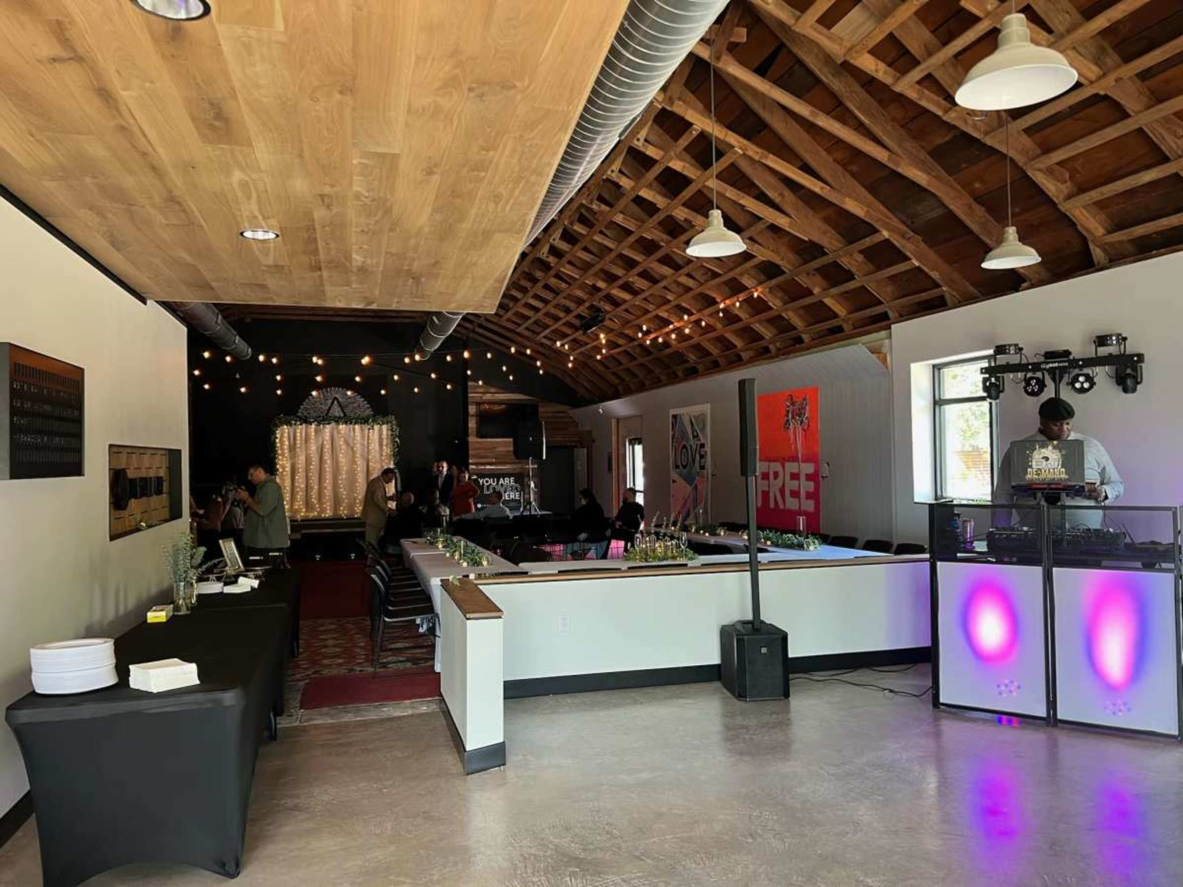 The image depicts a spacious indoor venue with a DJ setup, seating areas, and decorative lighting, surrounded by wooden beams and a modern aesthetic.