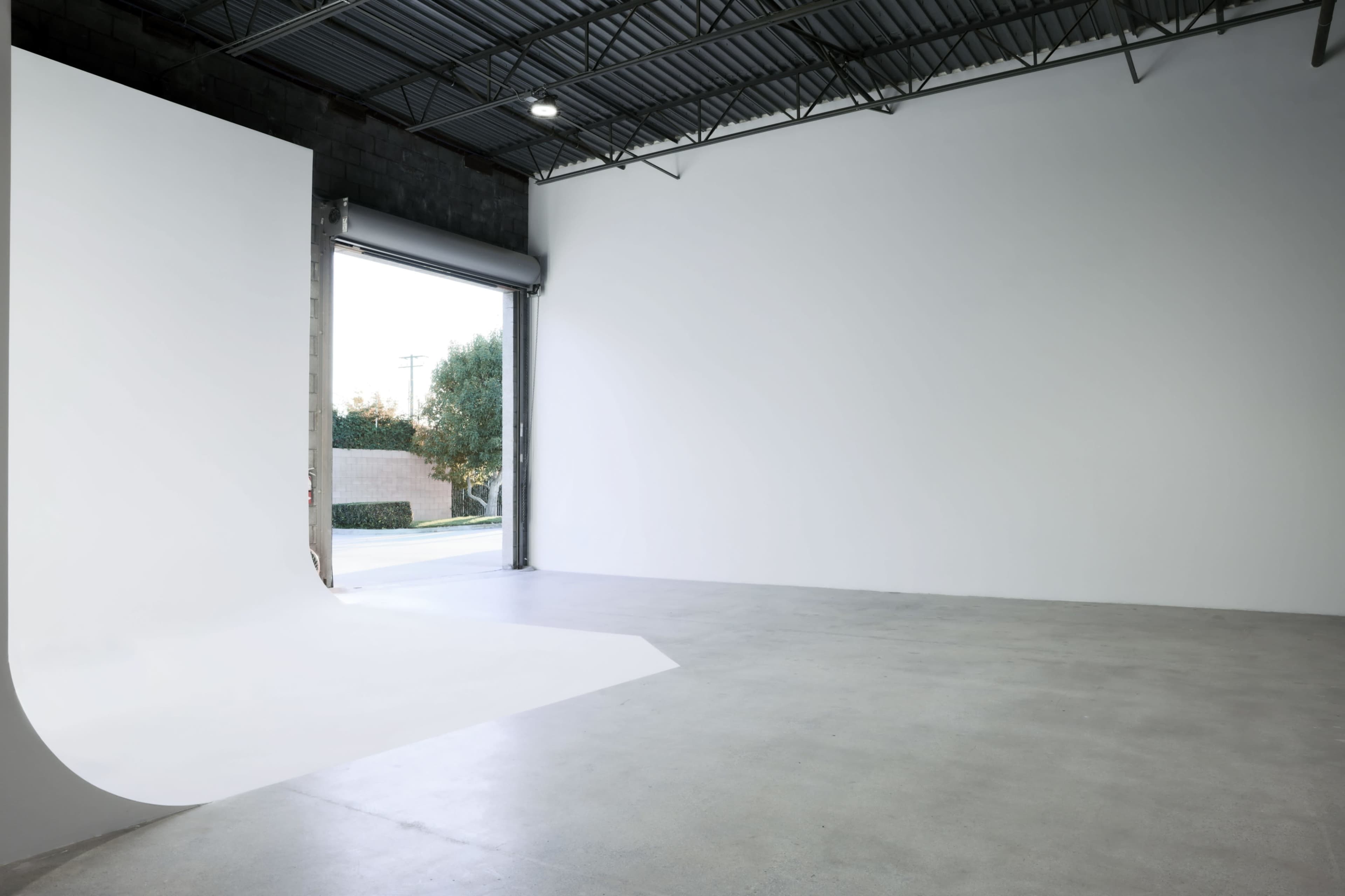 An empty indoor space features a large white backdrop and an open garage door leading to an outdoor area.