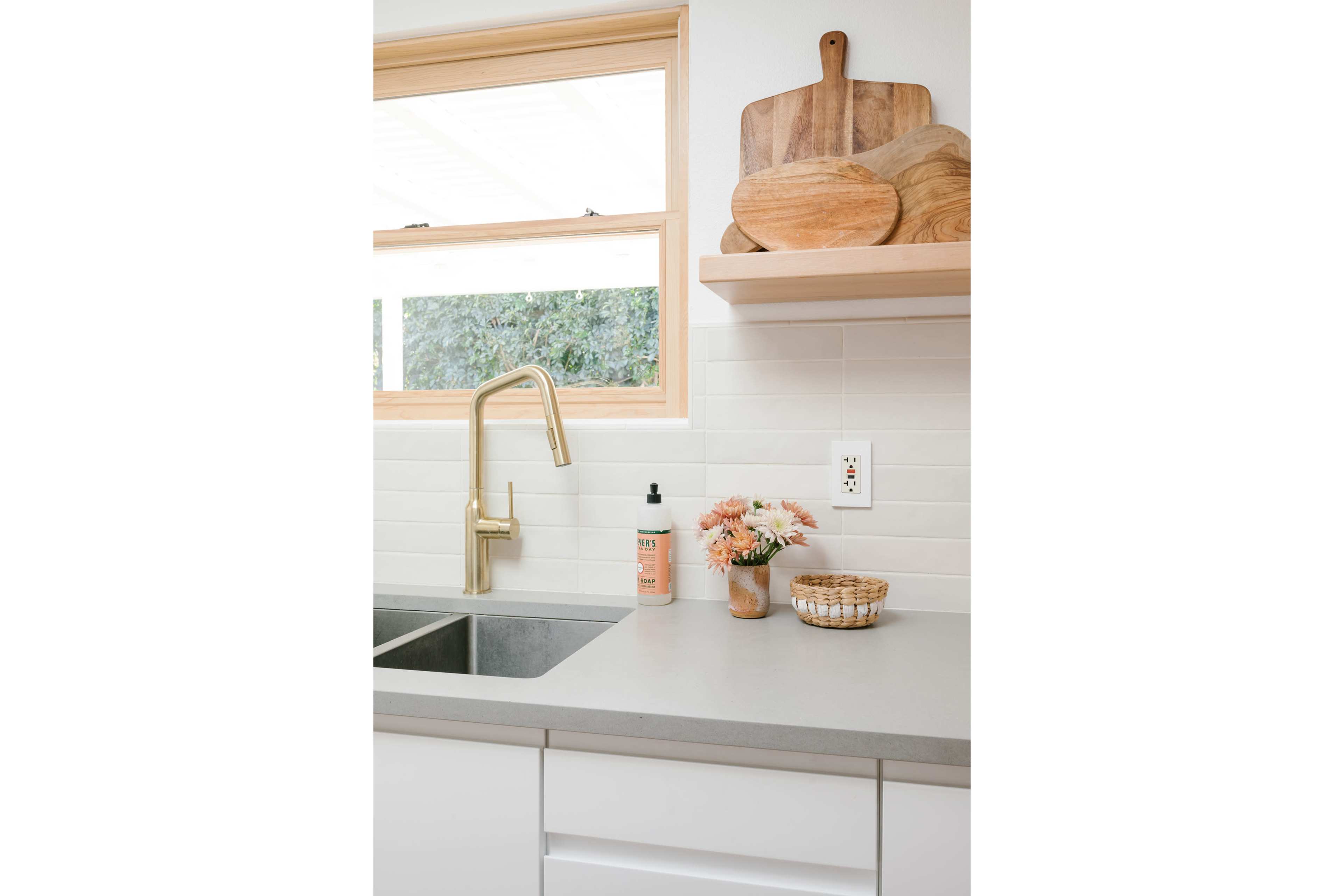 The image shows a clean kitchen sink area with a gold faucet, a small bouquet of flowers, and wooden cutting boards displayed above a light gray countertop.