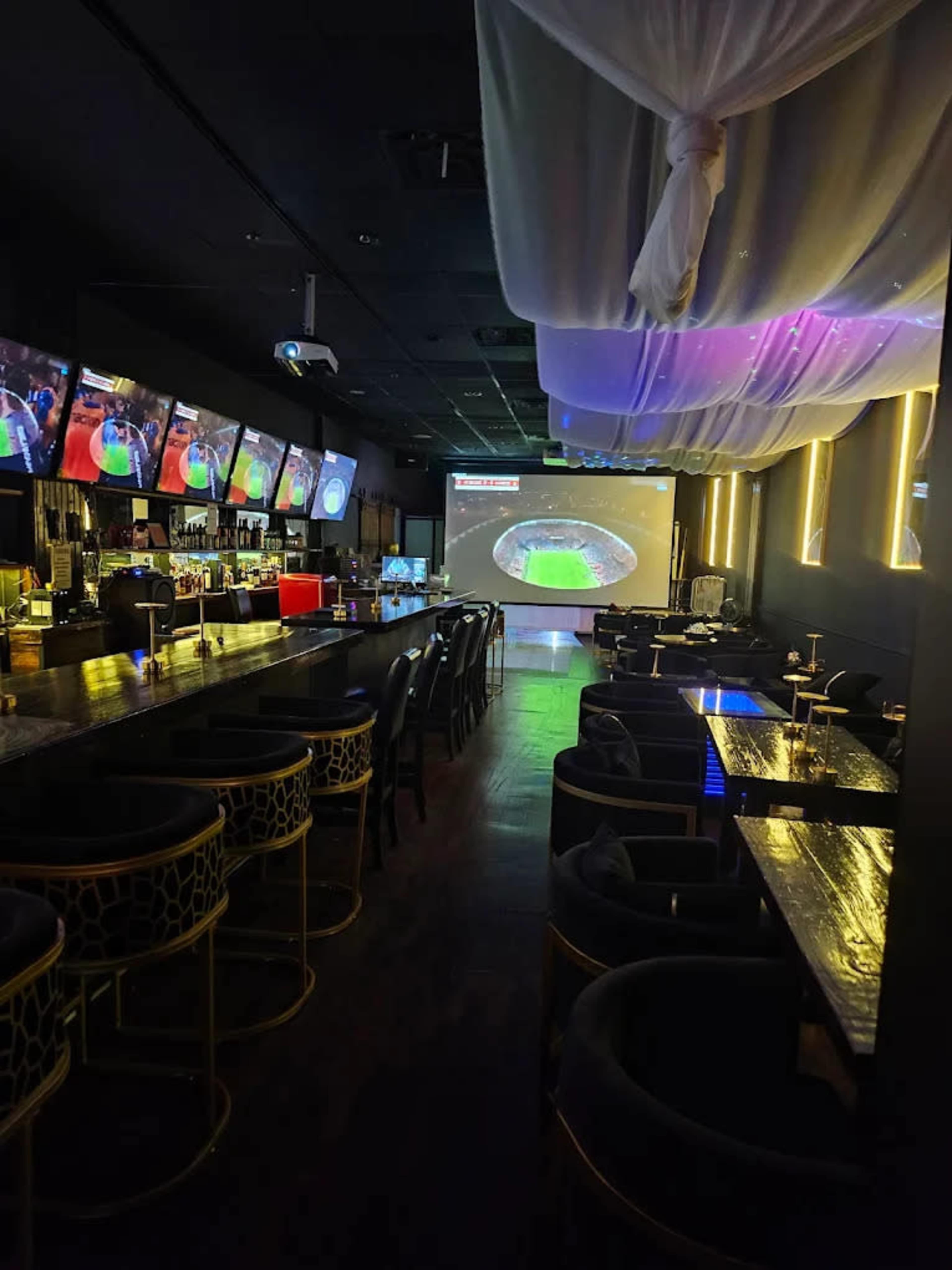 The image shows a modern bar interior with multiple screens displaying a sports game, a long bar counter, and lounge seating.