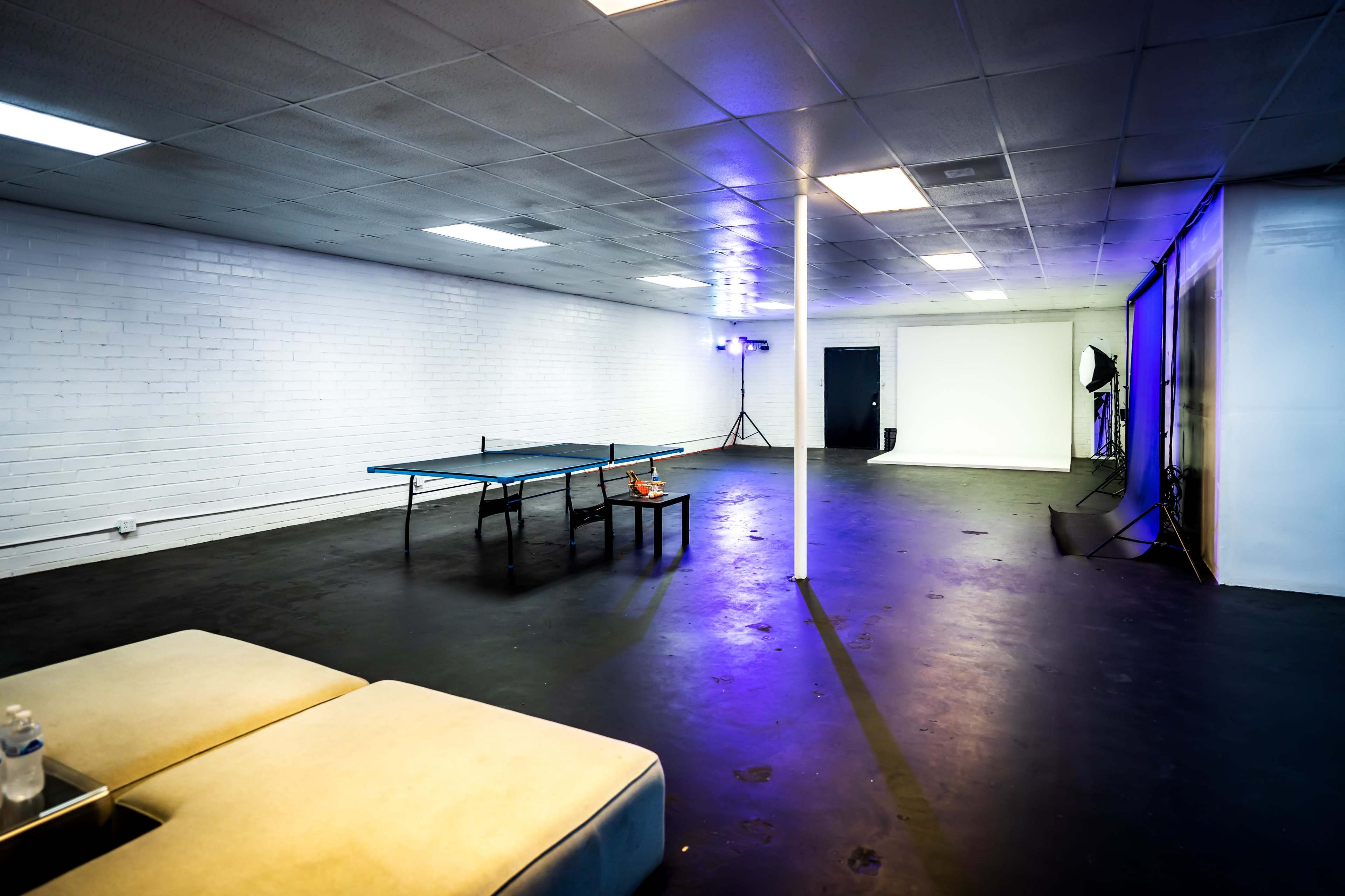 A spacious room with a ping pong table on one side, a small table with a drink, and a backdrop setup for photography.