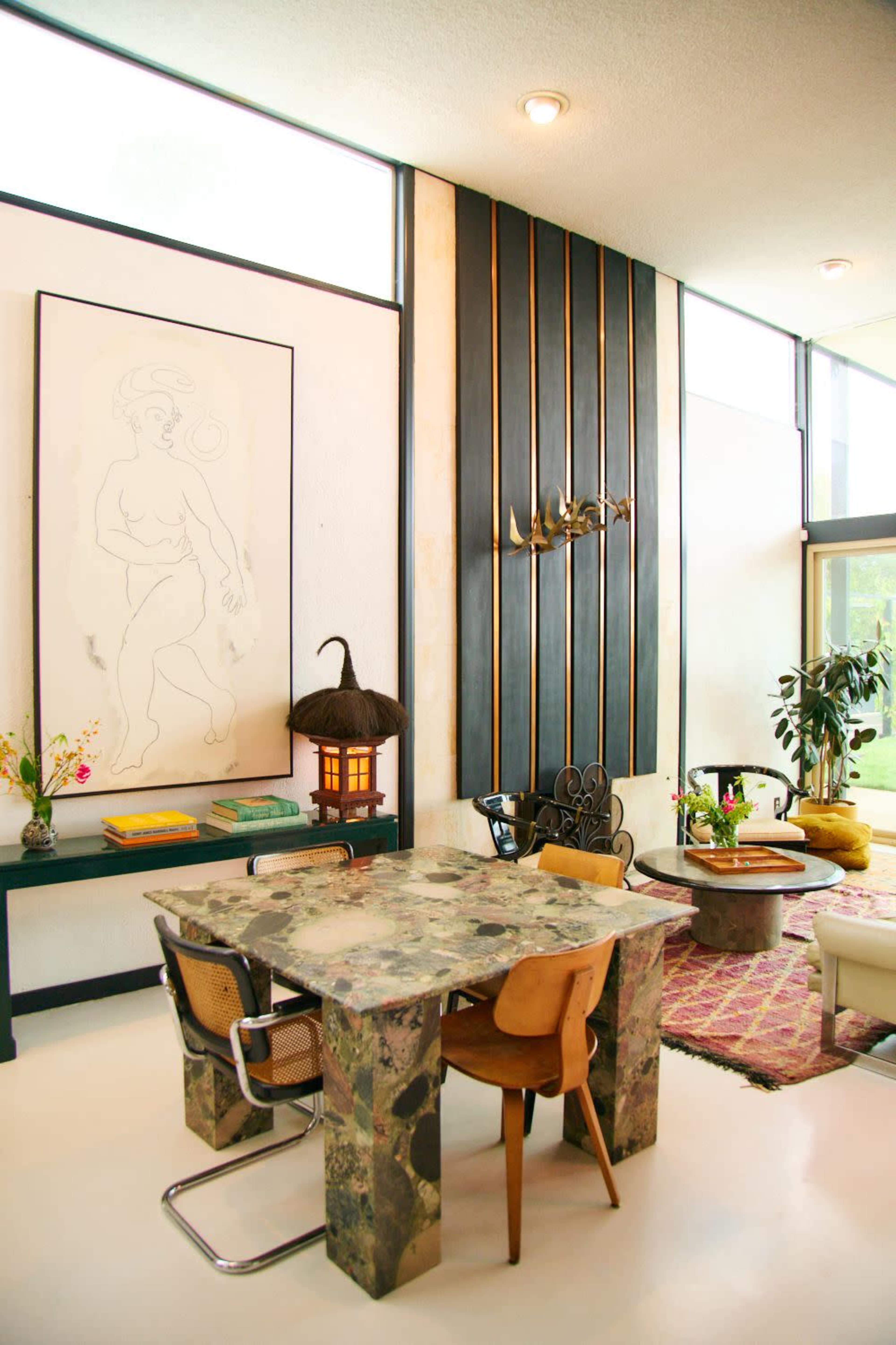 The image shows a modern interior space featuring a patterned stone dining table surrounded by various chairs, a large artwork on the wall, and a mix of decorative plants and furnishings.