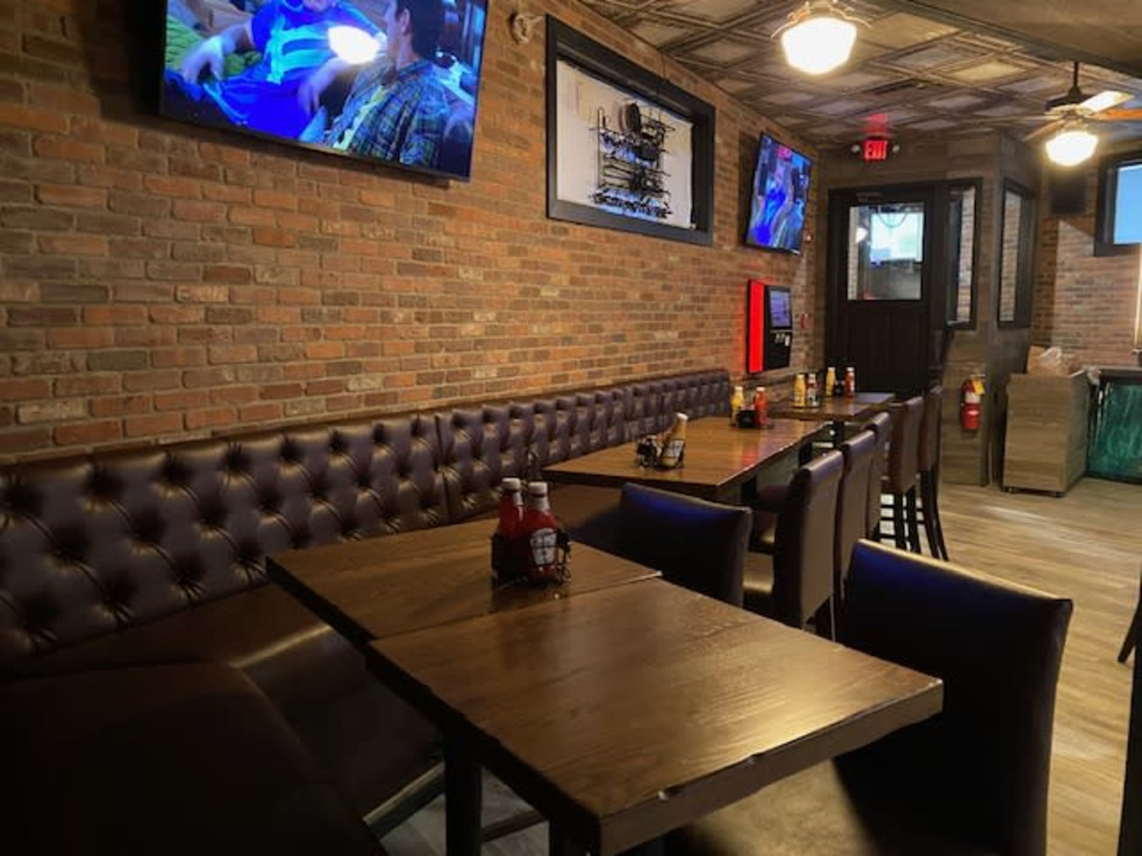 The image shows a casual dining area with wooden tables, upholstered seating, and multiple televisions mounted on the brick walls.