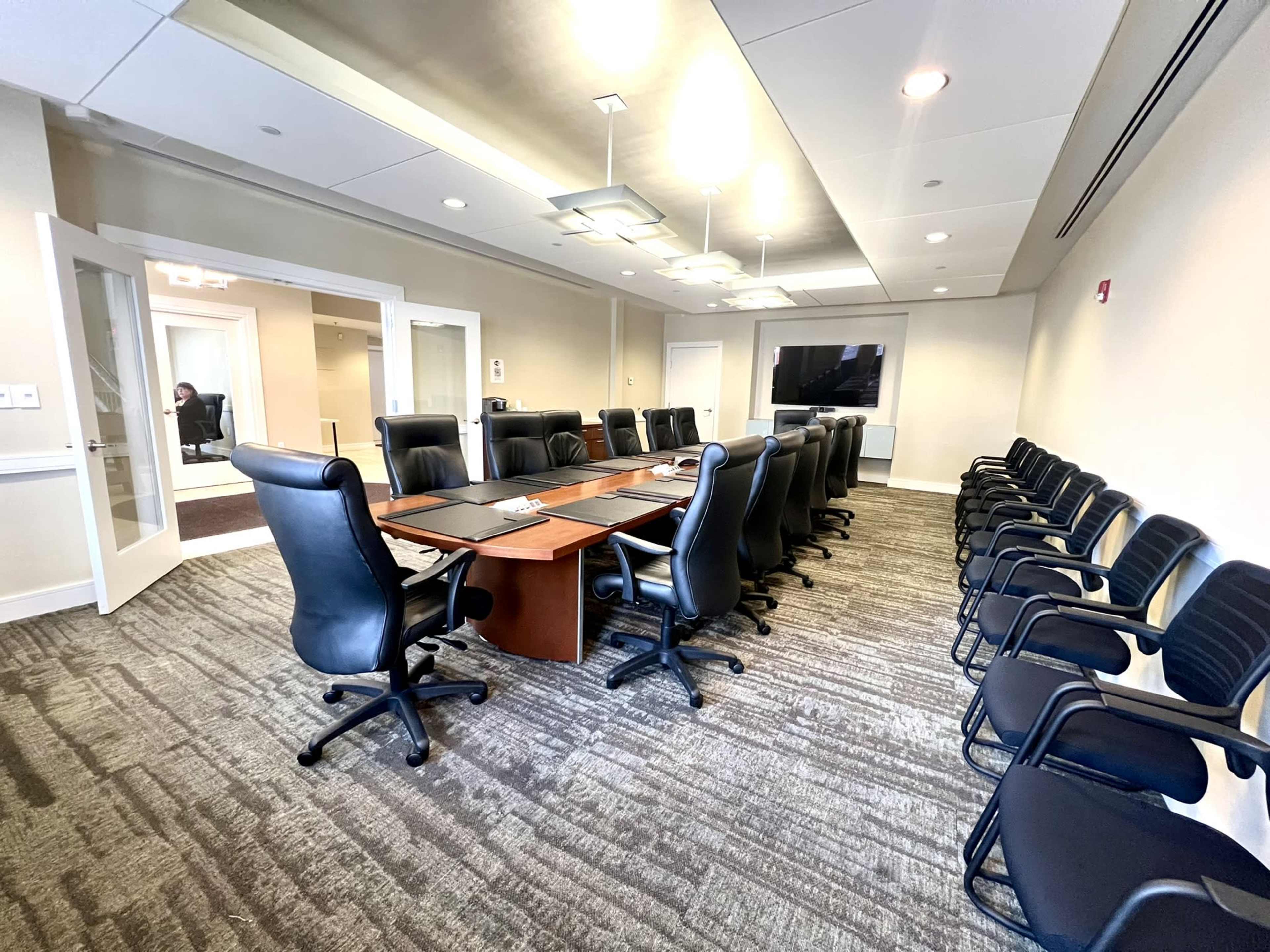 A large conference room features a long rectangular table surrounded by black leather chairs, with additional chairs aligned against the walls and a TV screen mounted at one end.