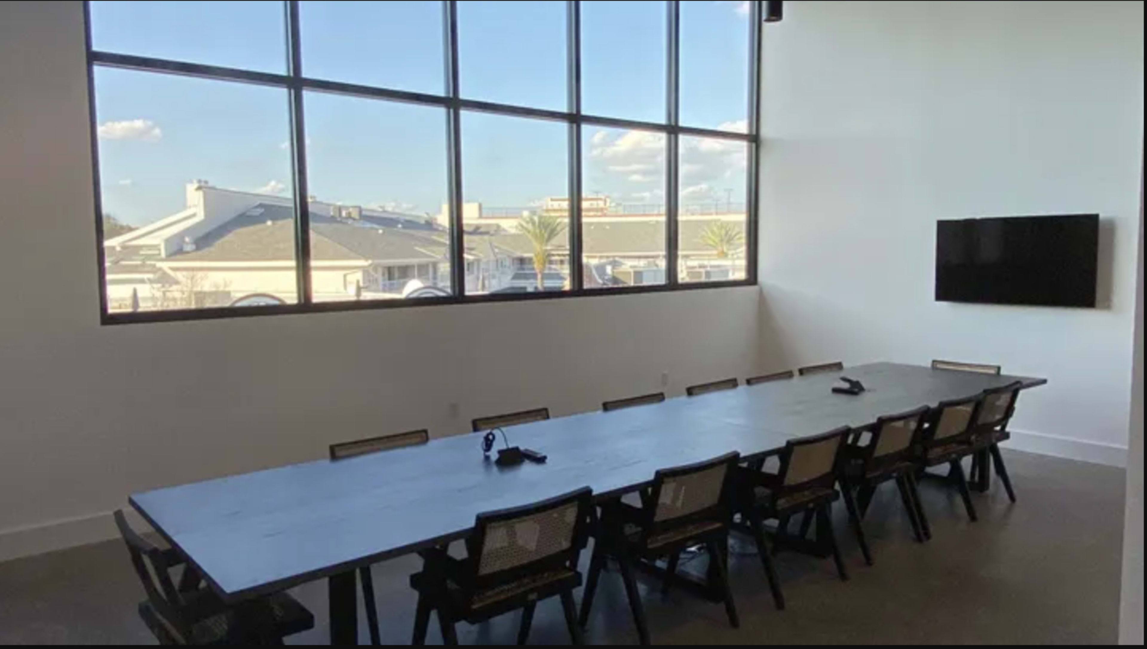 A spacious conference room features a long wooden table surrounded by chairs, large windows providing natural light, and a wall-mounted television.