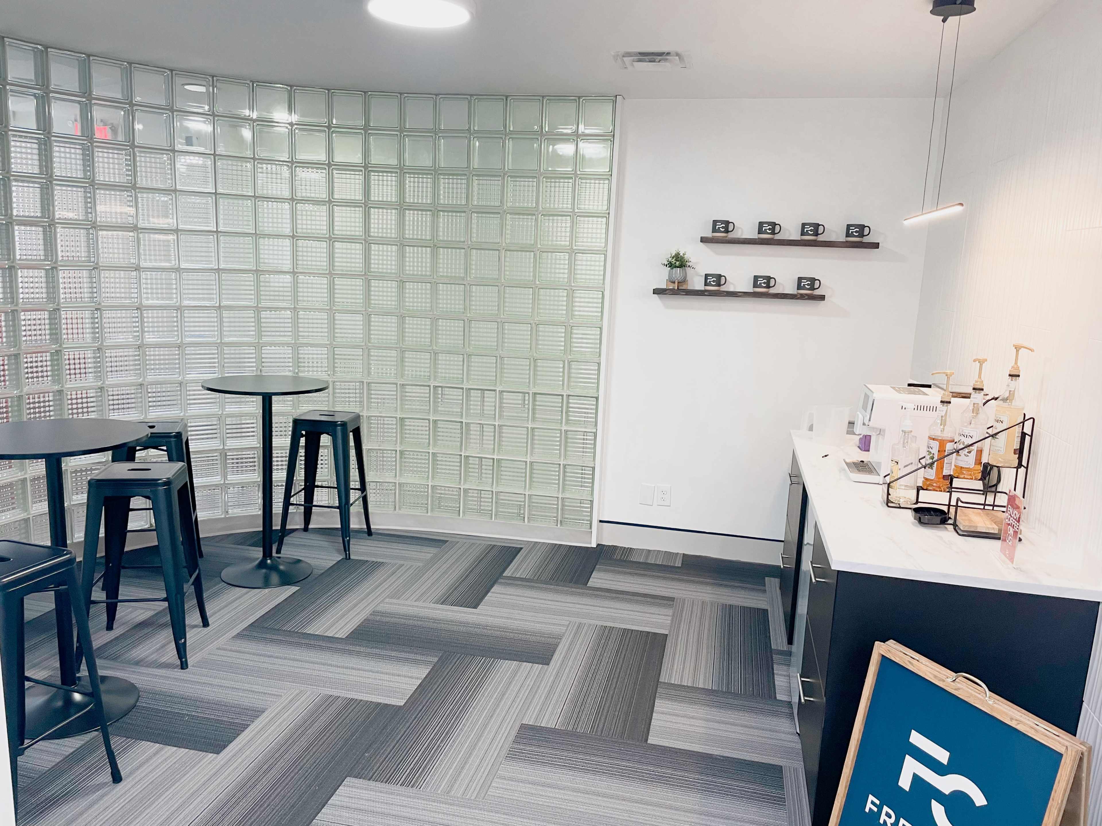 A modern break area featuring two high tables with stools, a coffee station, and a glass block wall.
