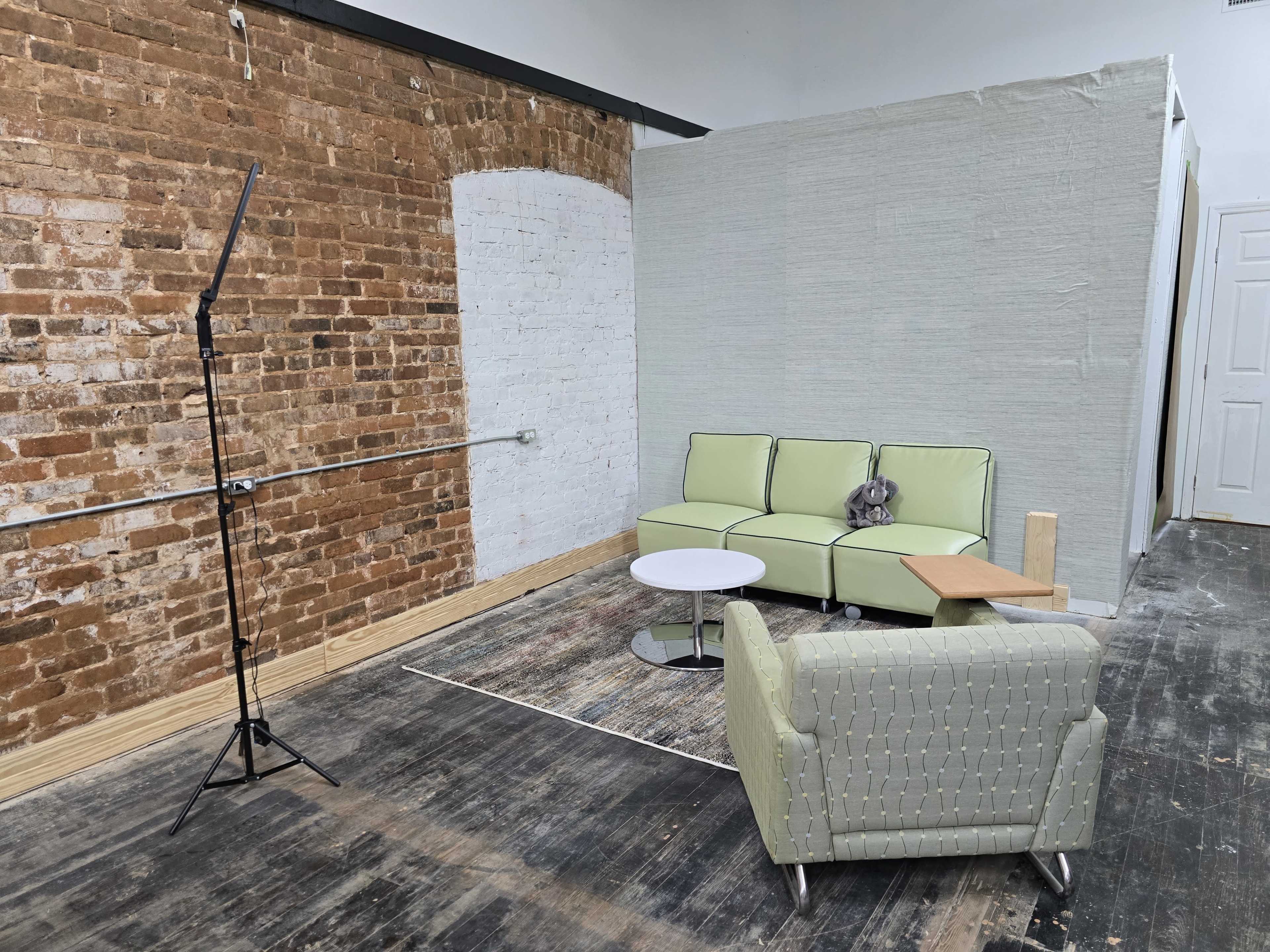 The image shows a minimalist interior space with a light green sectional sofa, a white coffee table, a patterned armchair, and a standing lamp, all set against exposed brick walls.
