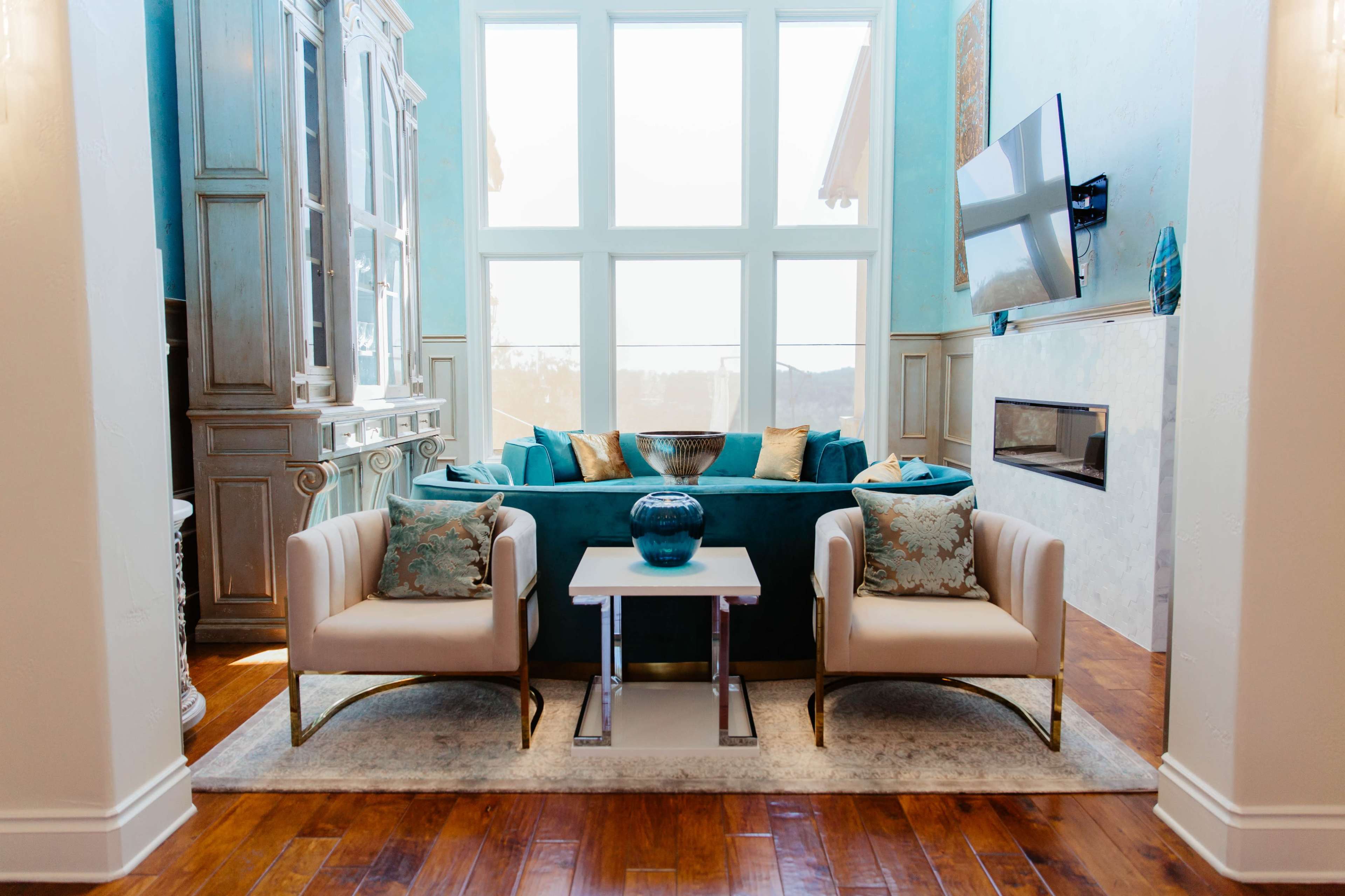 A modern living room featuring a turquoise couch, two white chairs, a glass coffee table, and large windows letting in natural light.