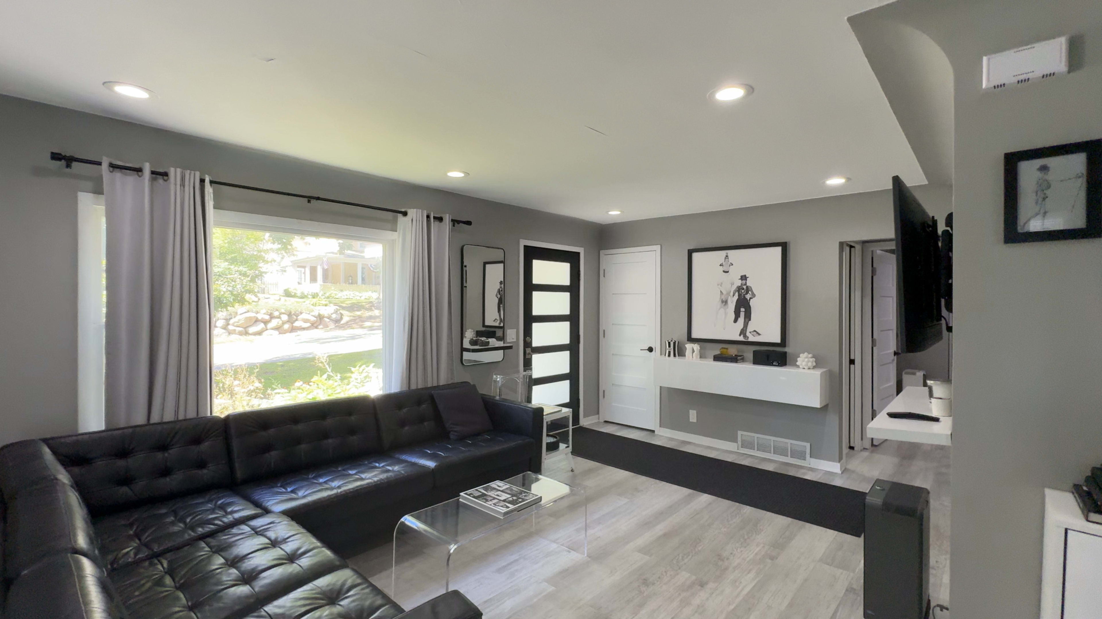 The image shows a modern living room with gray walls, a black sectional sofa, and a large window letting in natural light.