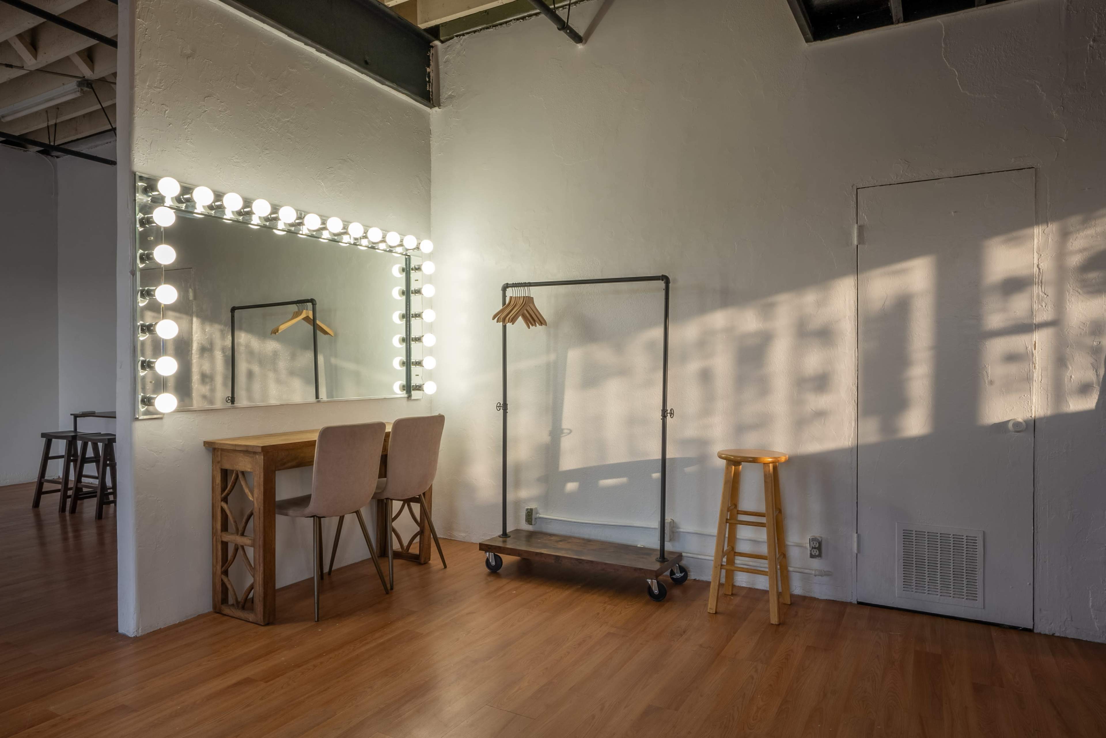 The image features a minimalist interior with a vanity mirror surrounded by lights, a small table, a clothing rack on wheels, and a wooden stool, all set against a wall with soft shadows.