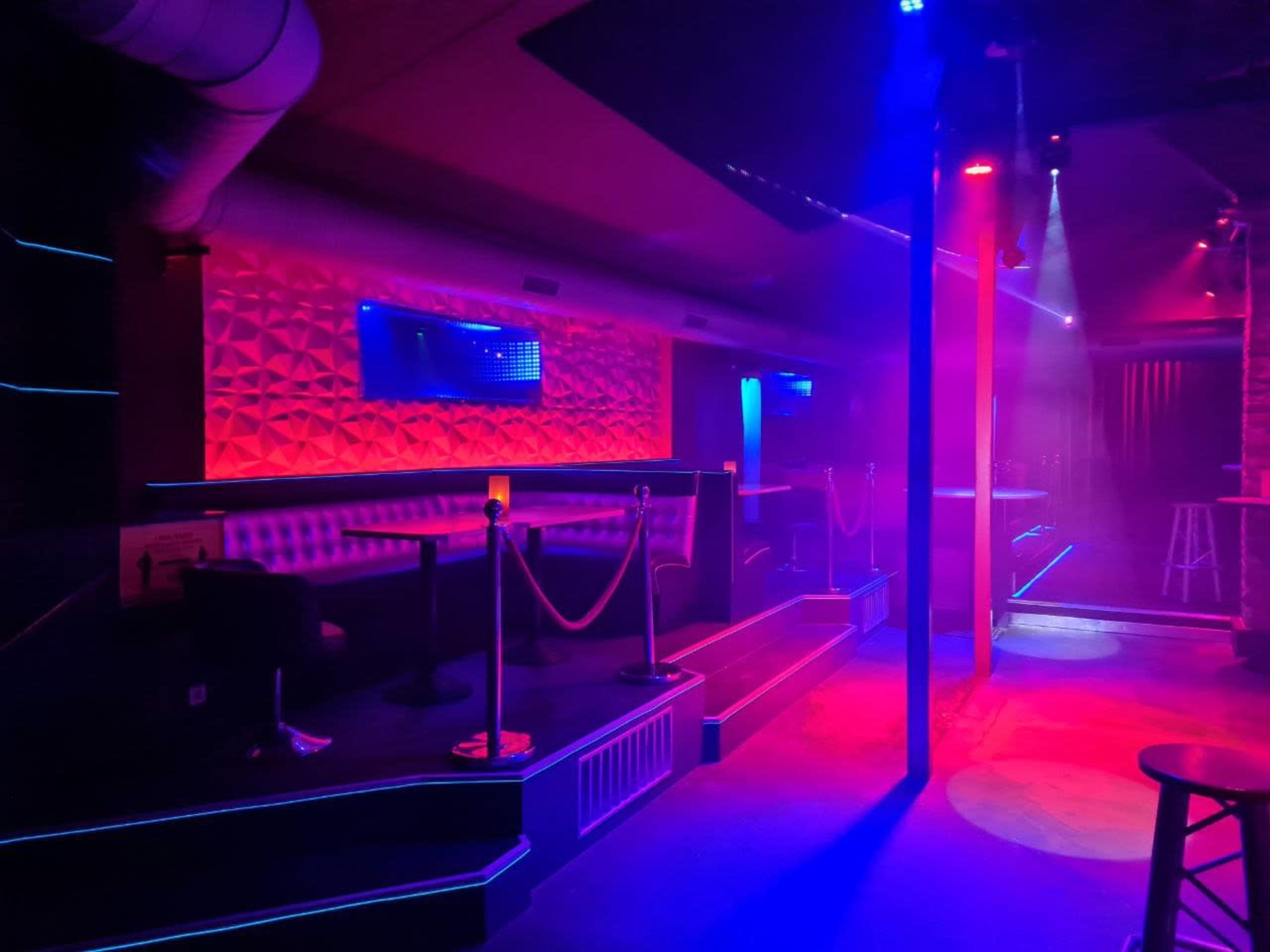 A dimly lit bar interior with blue and red lights, featuring seating areas separated by ropes and empty space illuminated by stage lights.