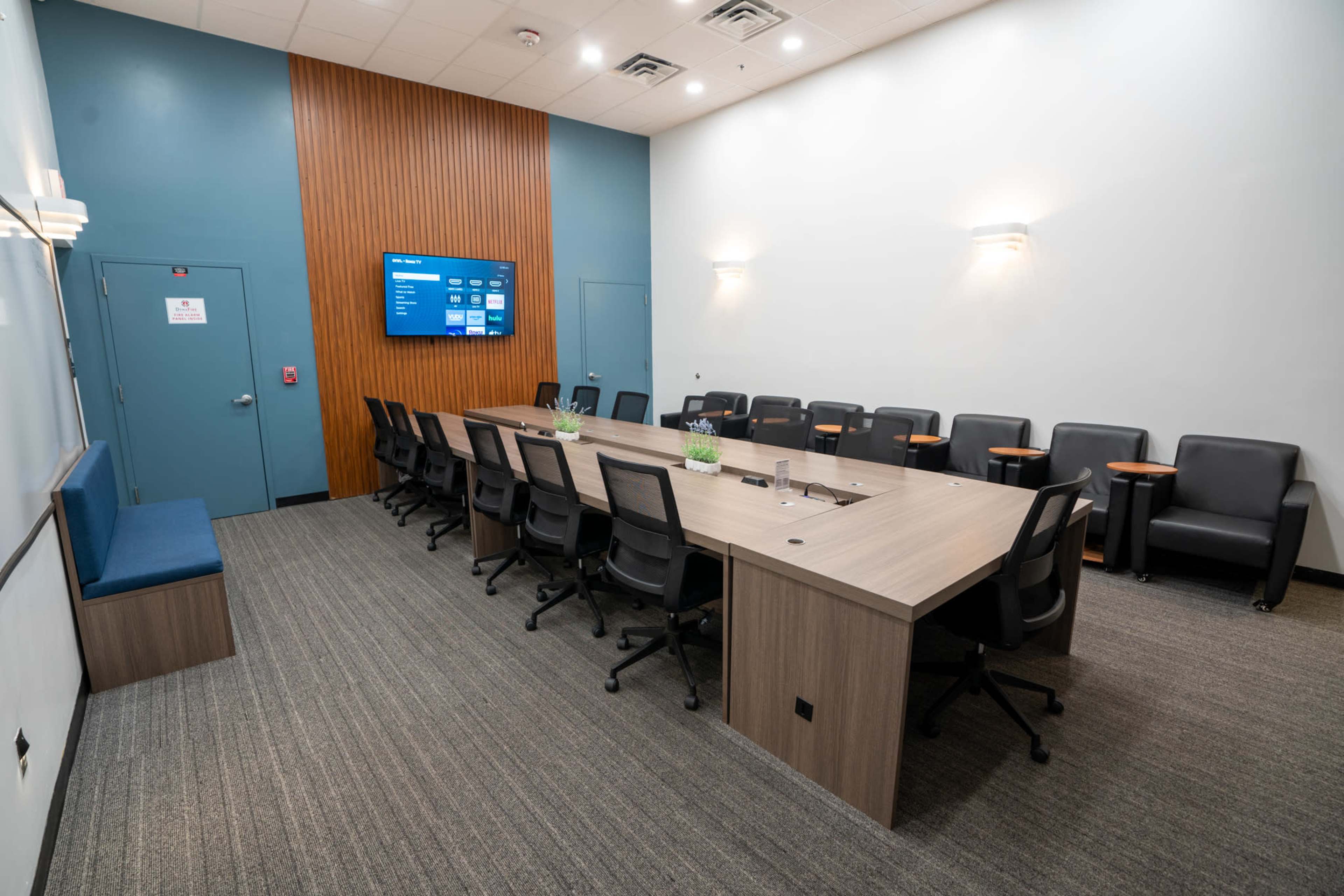 A modern conference room with a large table, multiple black chairs, and a wall-mounted screen.